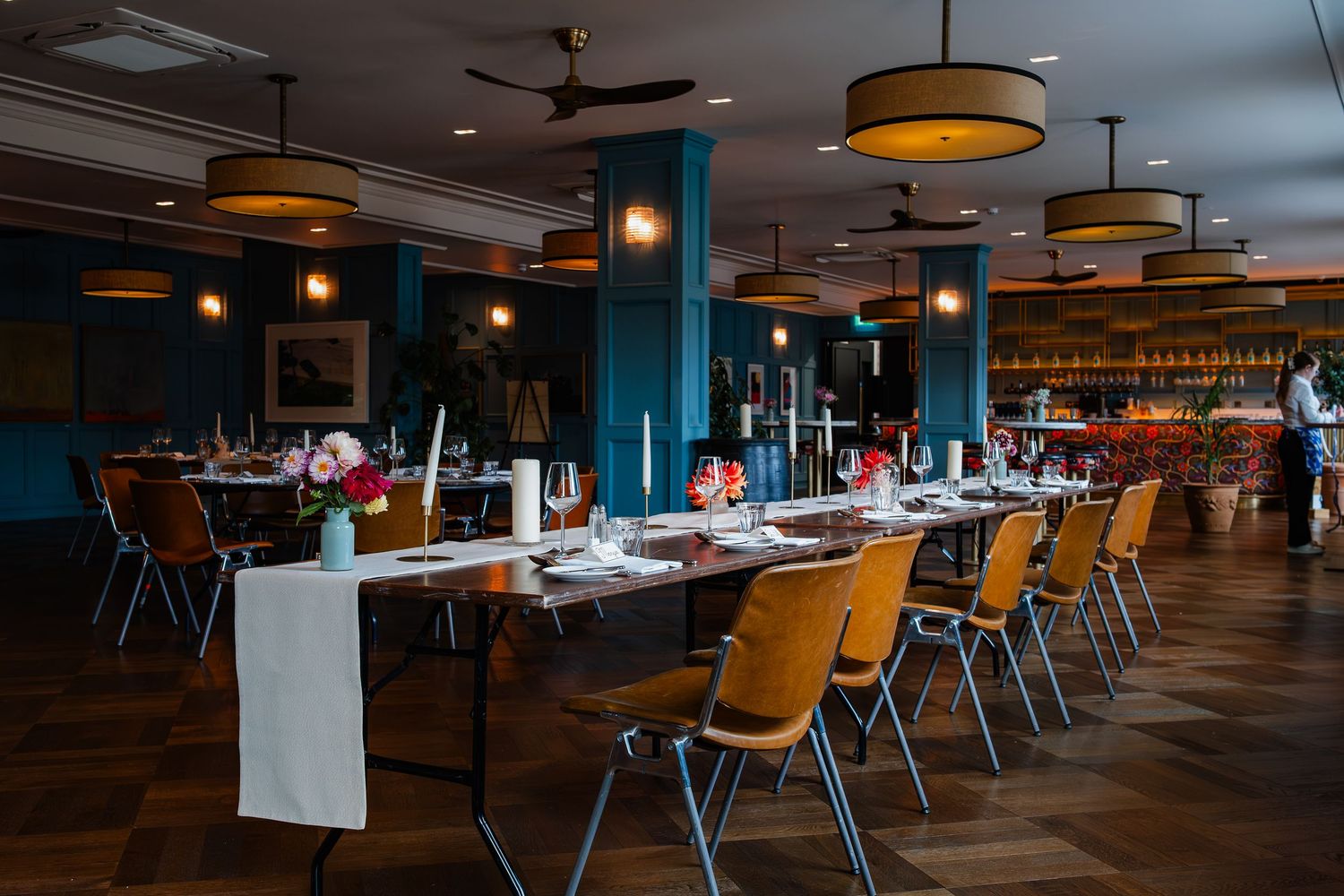 Modern restaurant interior with wooden tables and chairs arranged in rows under pendant lighting fixtures.
