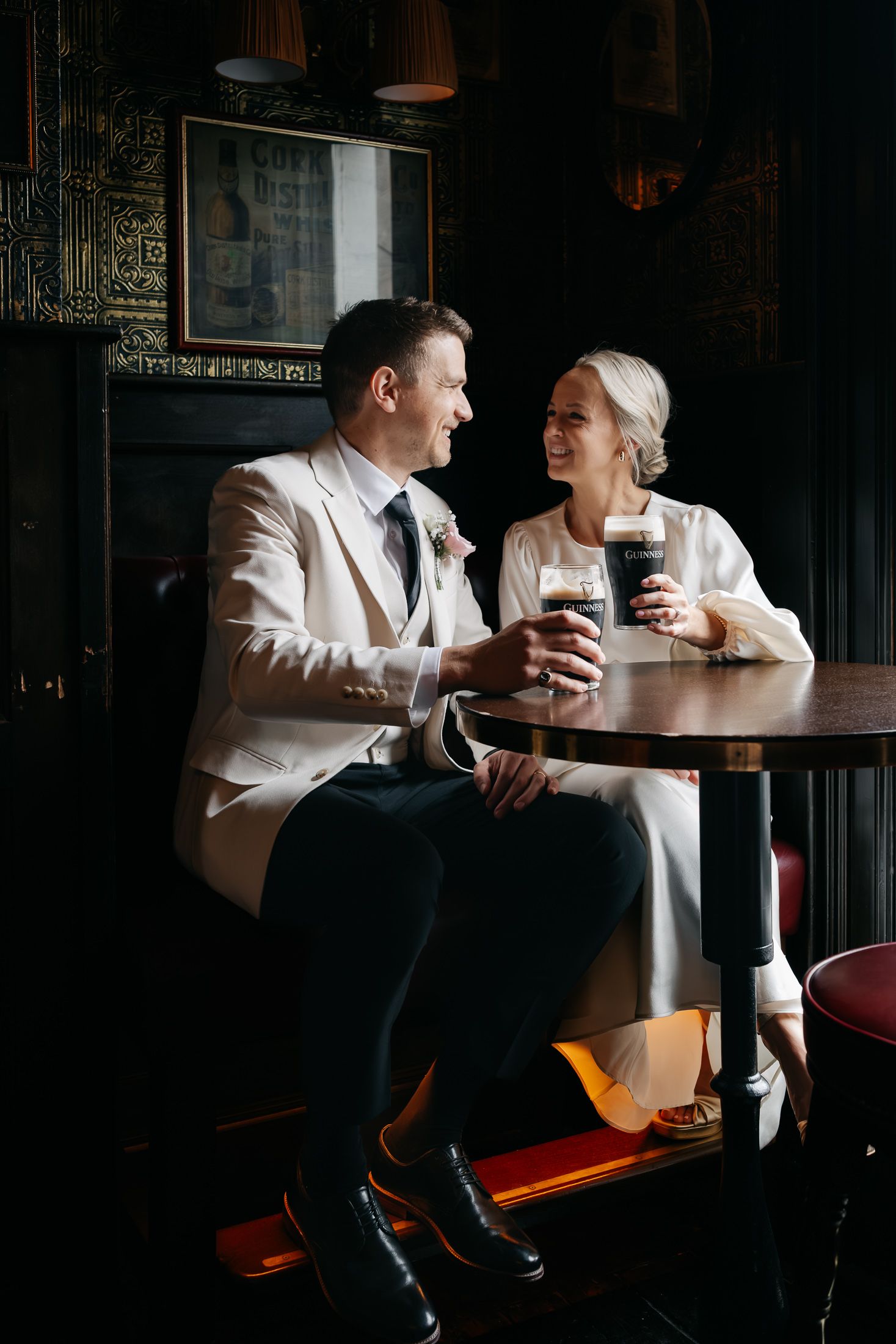 Bride and groom in wedding attire share Guinness pints at a dark, cozy pub, smiling at each other.