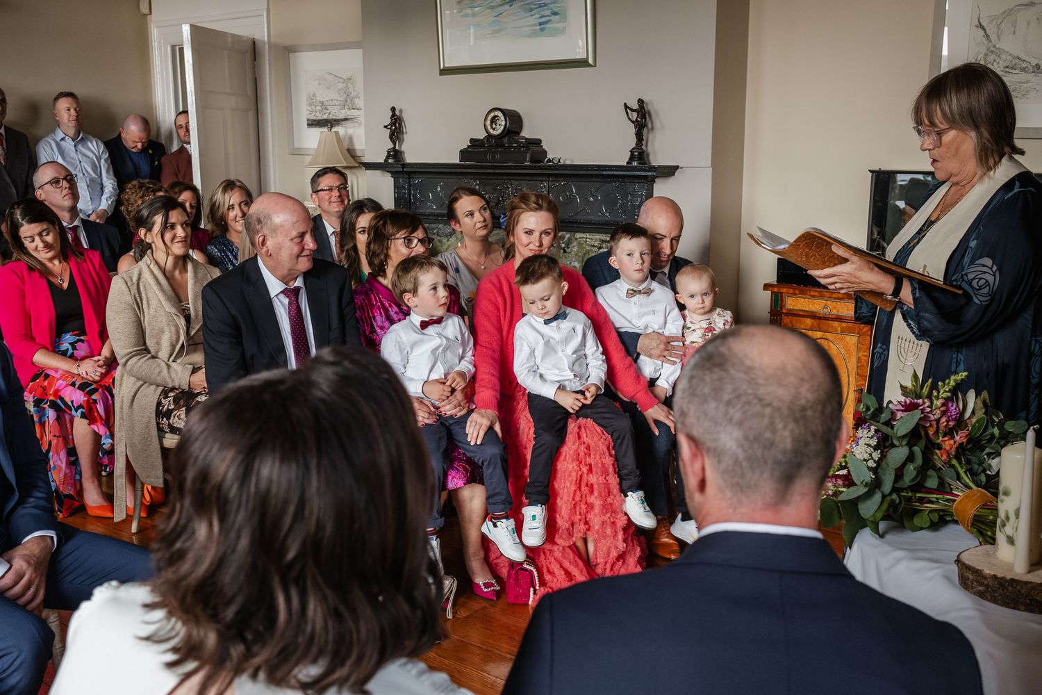 Family gathering indoors for a christening ceremony, with guests seated around babies in white outfits near a fireplace.