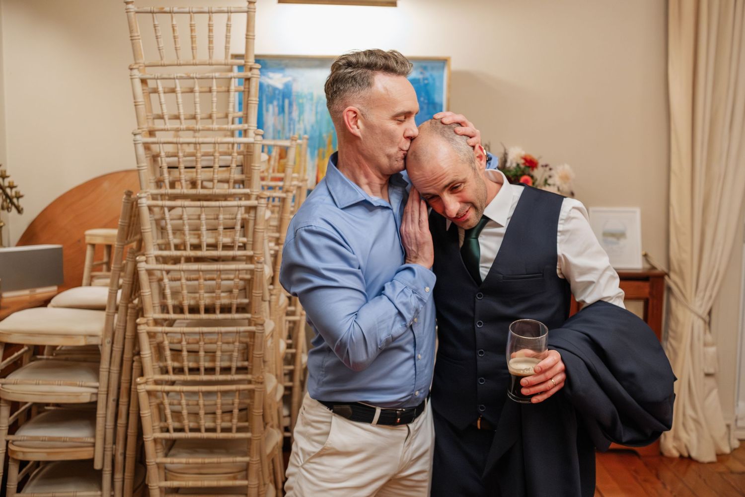 Two grooms share an intimate moment at their wedding reception, surrounded by stacked chiavari chairs.