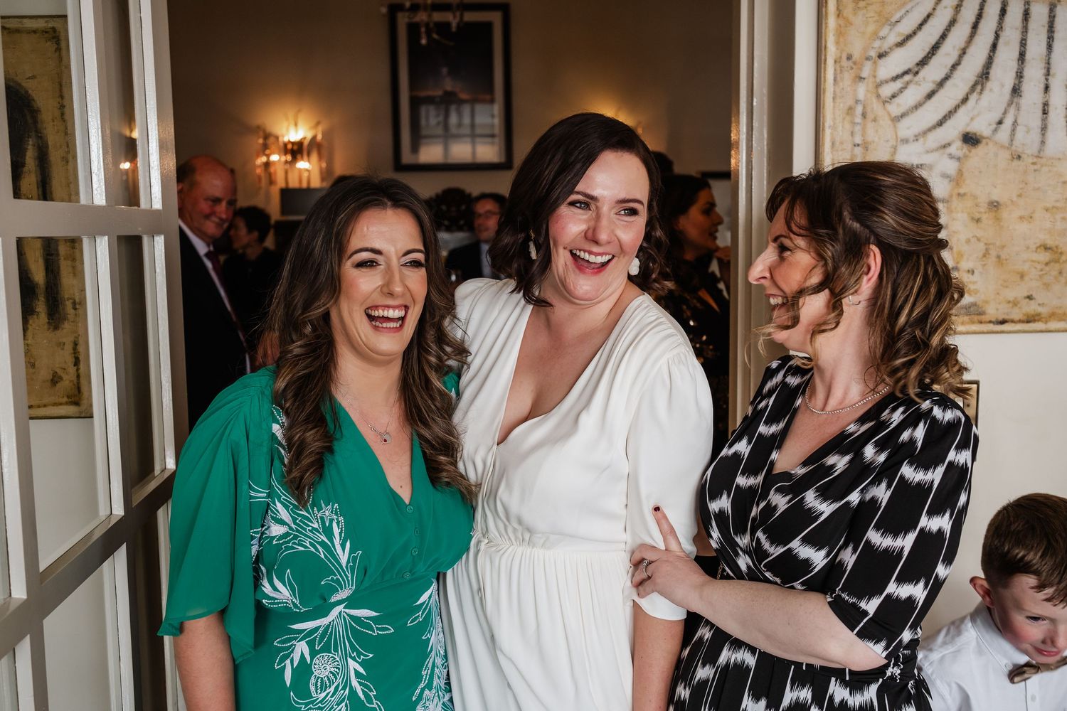 Three women smiling and laughing together at a wedding reception near a doorway.