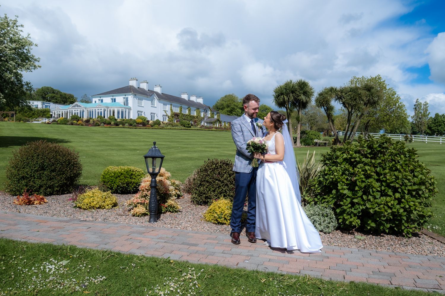 Bride and groom pose in lush garden grounds of elegant country house wedding venue on a sunny day.