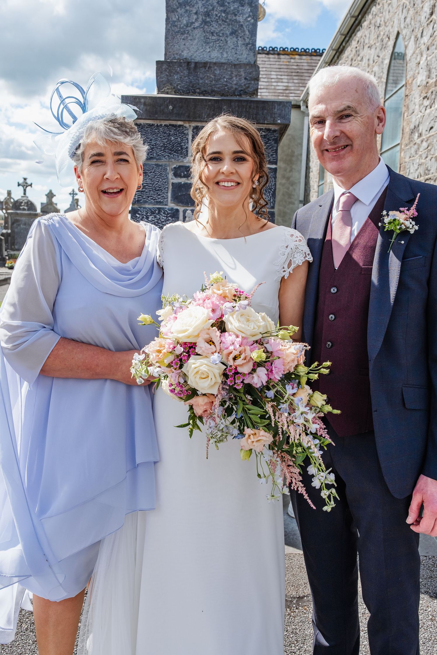 Wedding party in light blue and burgundy attire pose with pink floral arrangements outdoors.