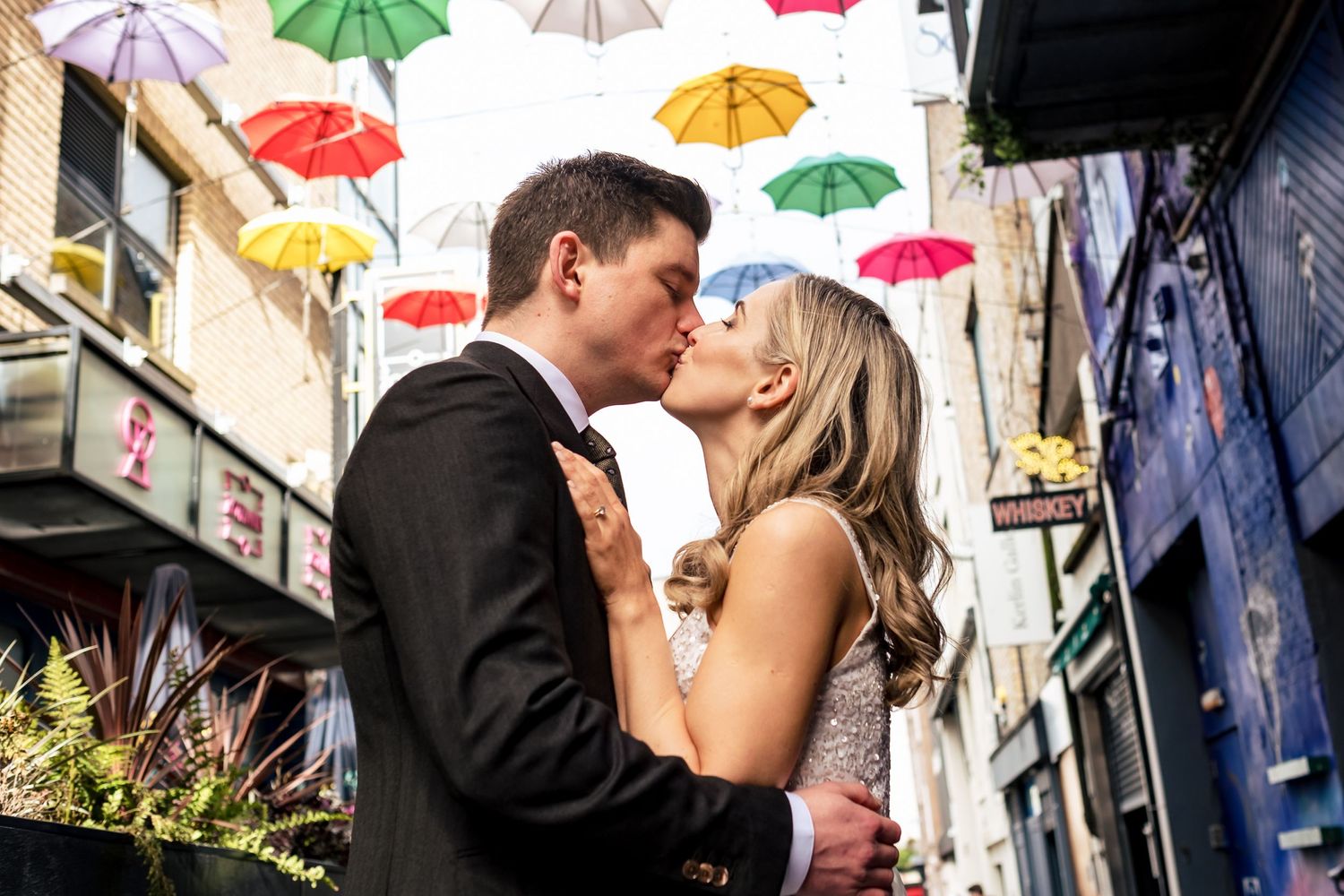 Couple kisses beneath colorful suspended umbrellas in urban alleyway.