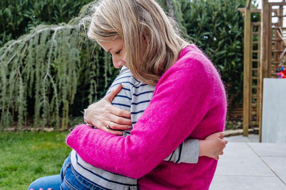 Woman in pink cardigan hugging a child outdoors in a garden setting.