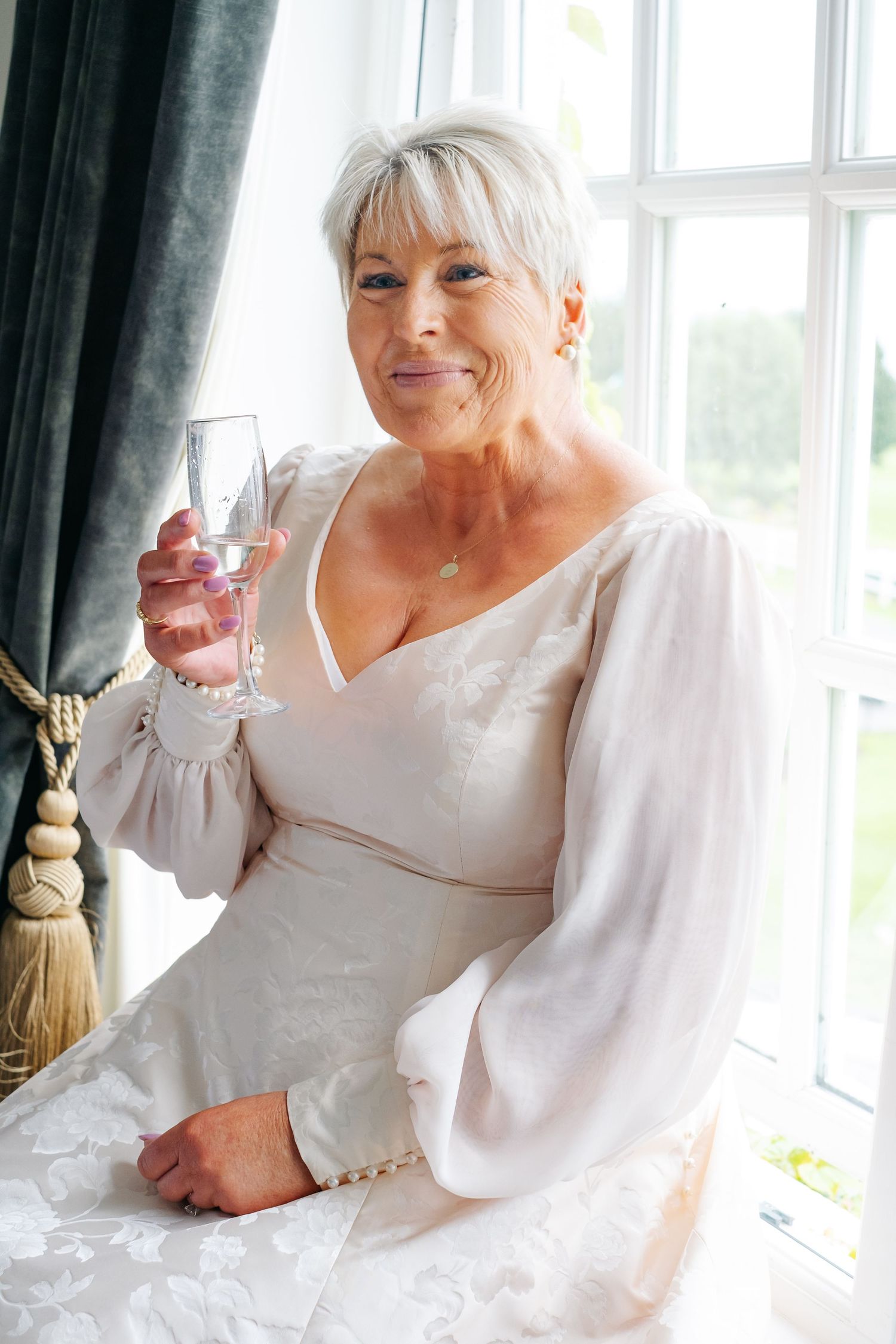 Elegant mature bride in white dress holding champagne glass by window on wedding day.