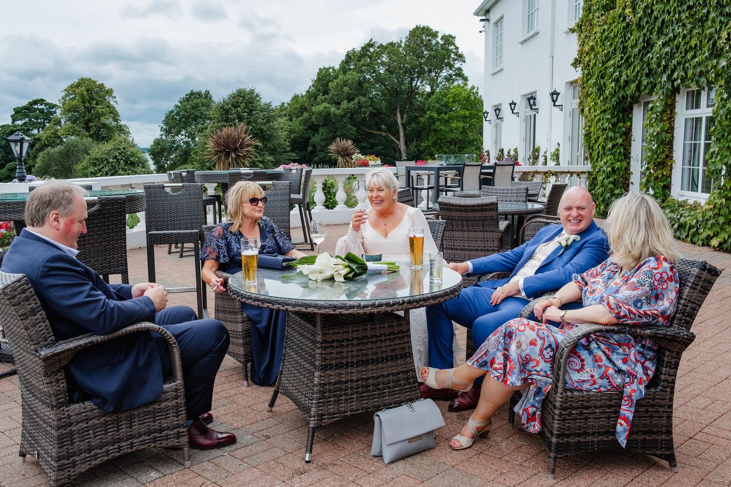 Five wedding guests enjoy drinks and conversation at an outdoor patio table surrounded by lush greenery.