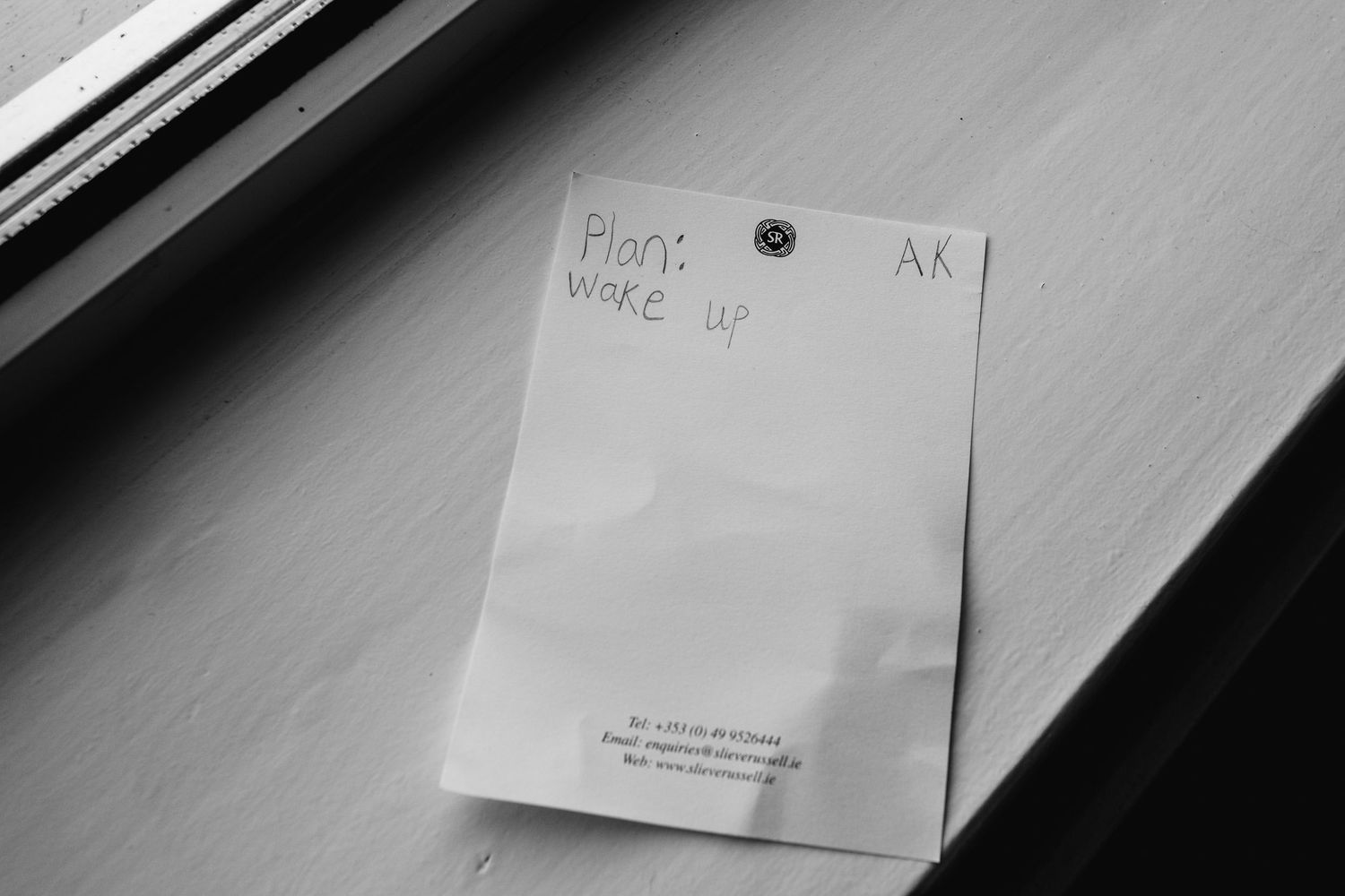 Black and white photo of a sticky note on a wooden surface reading Plan: Wake Up with initials AK.
