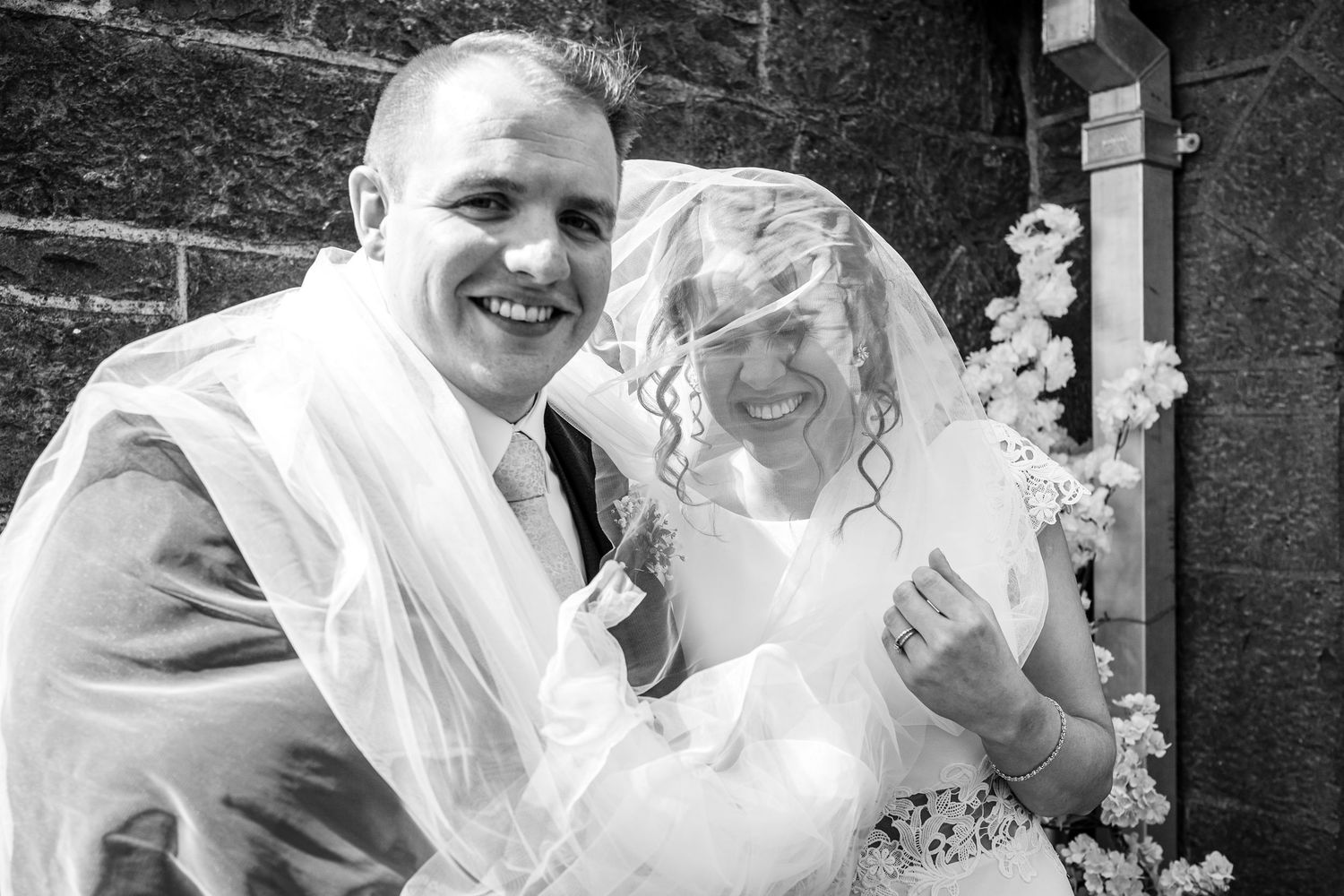 Black and white wedding photo of smiling bride and groom, veil blowing in wind outside stone church.