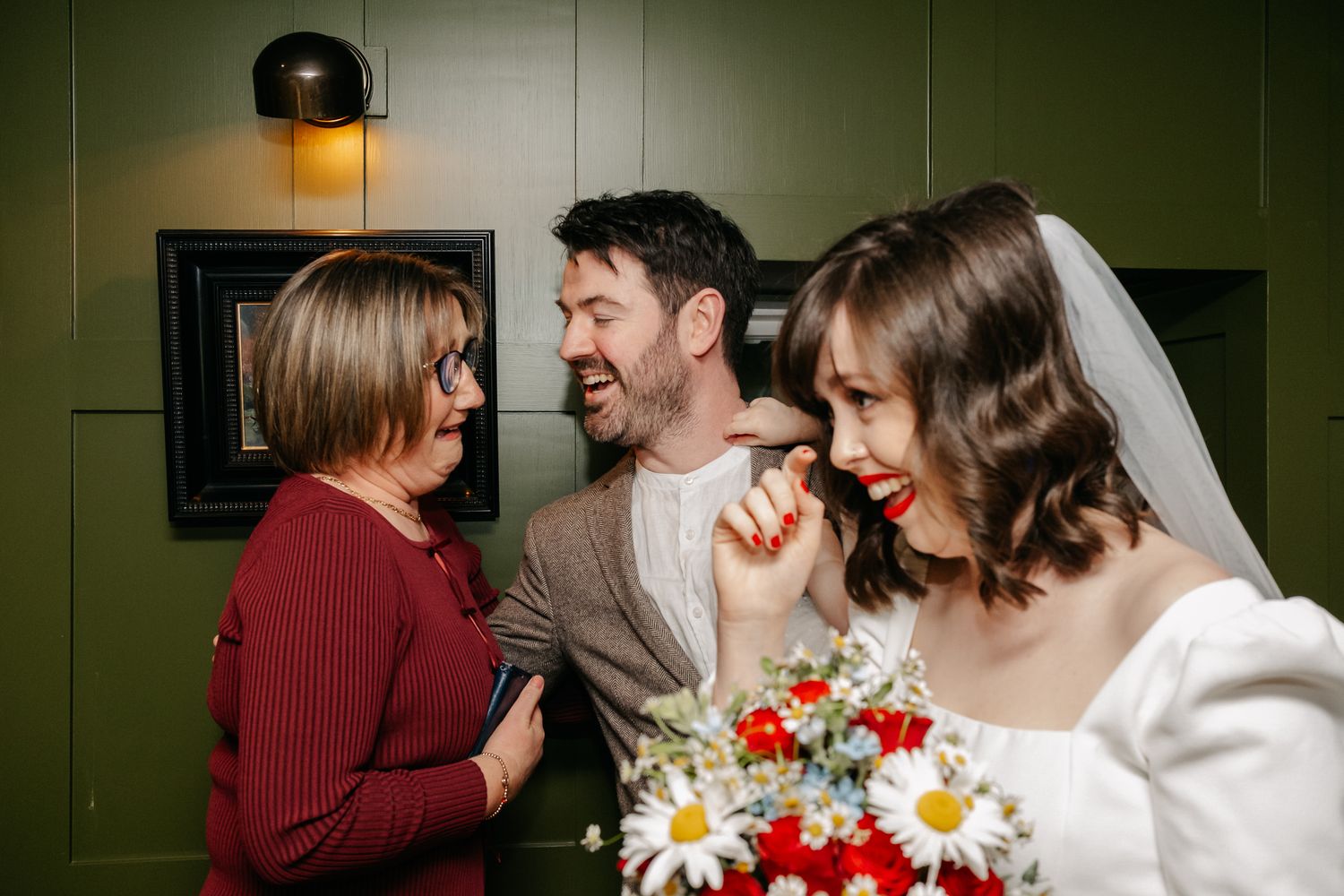Bride in white dress and veil laughing with groom and female guest at wedding reception.