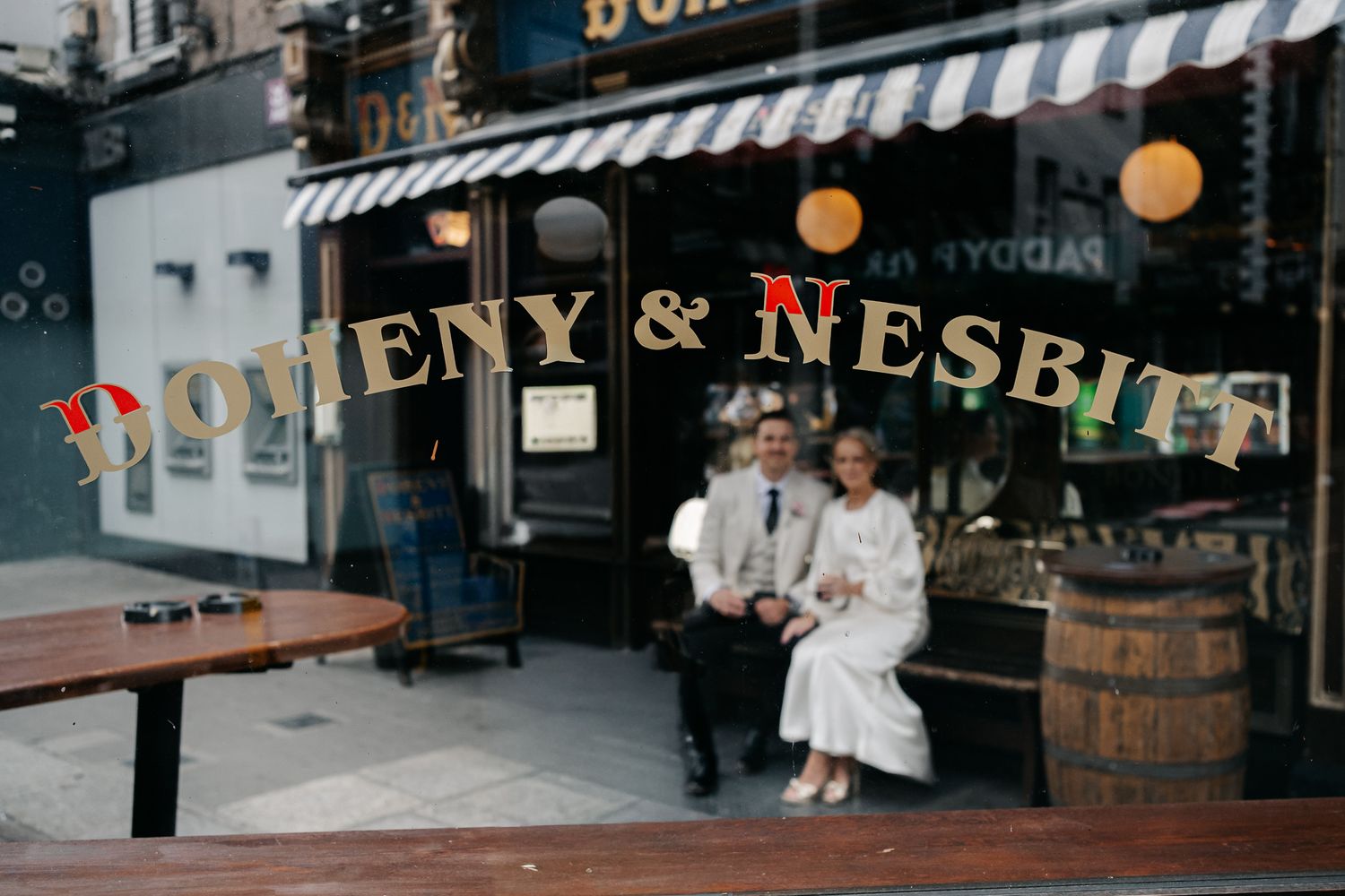 Couple sits outside Boheny & Nesbitt pub under colorful banner, woman in white dress, man in light suit.
