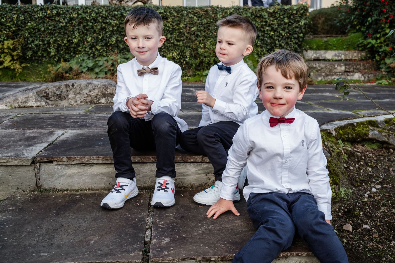 Three young boys in white shirts and bow ties sit on stone steps, dressed smartly for a formal occasion.
