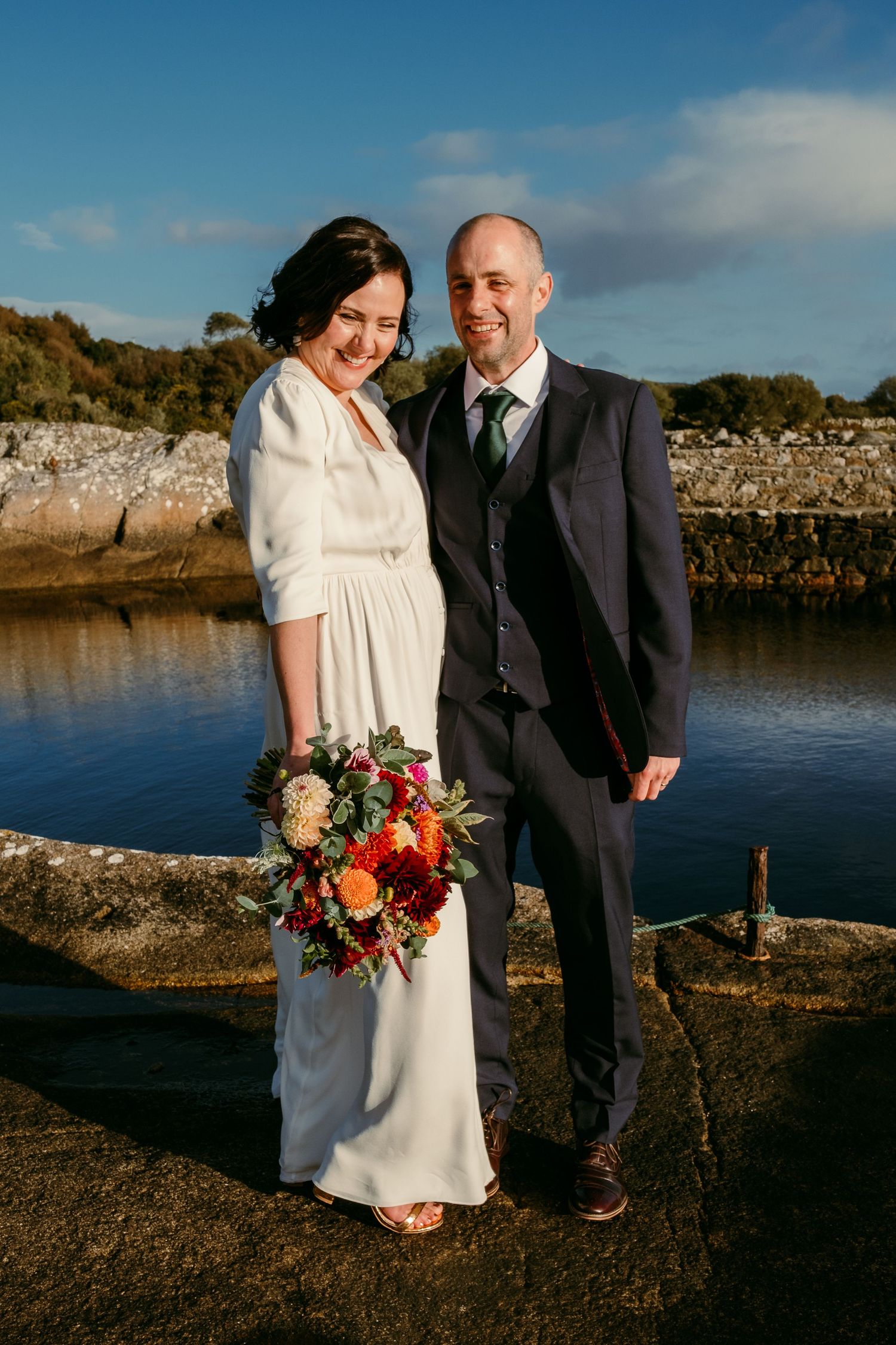 Bride and groom pose together at a waterfront wedding venue, bride holding a vibrant autumn bouquet.