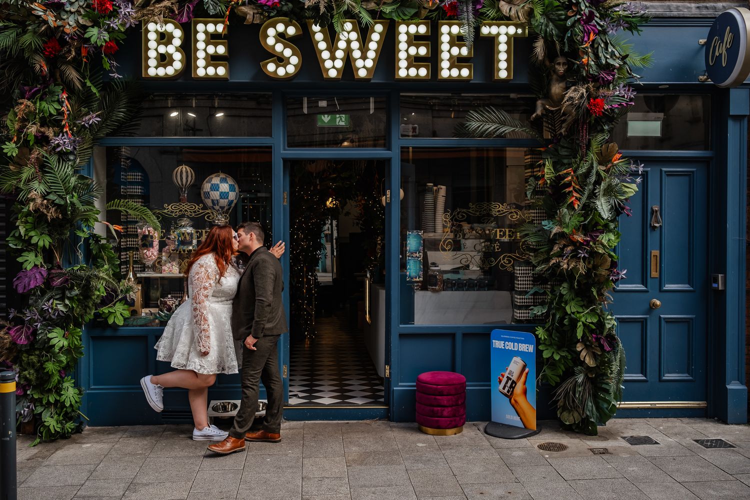 Illuminated 'BE SWEET' sign decorates a storefront with festive holiday greenery.