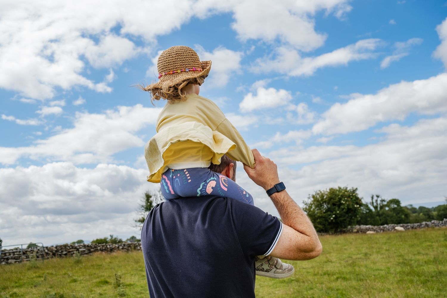 Person carrying child on shoulders during sunny day in countryside.