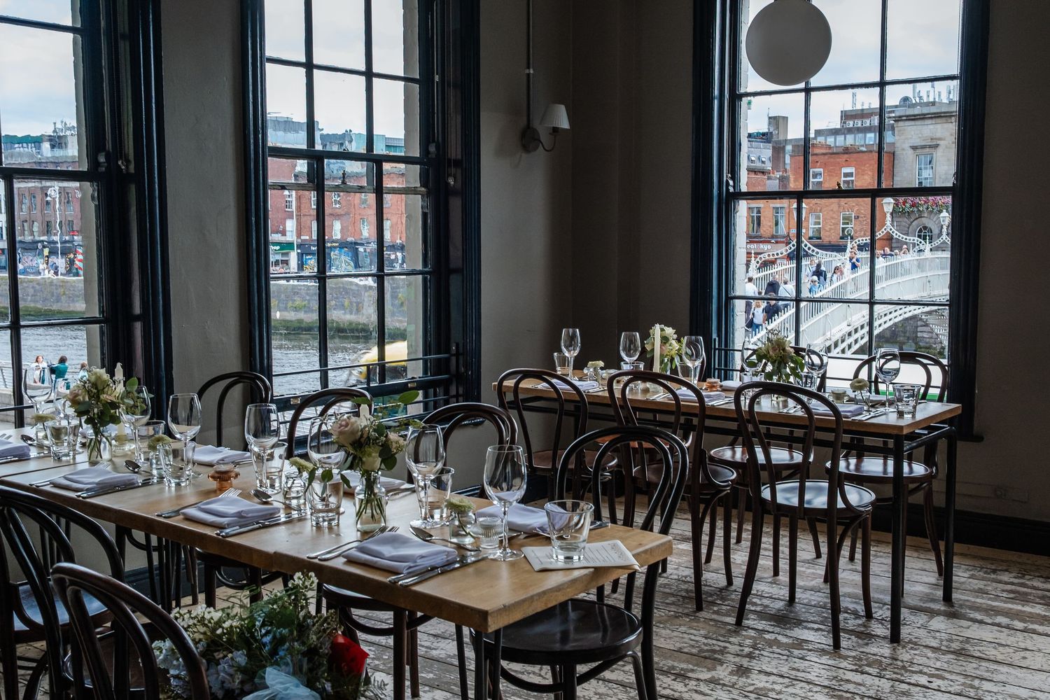Modern restaurant dining room with floor-to-ceiling windows, wooden tables, and black chairs overlooking city buildings.