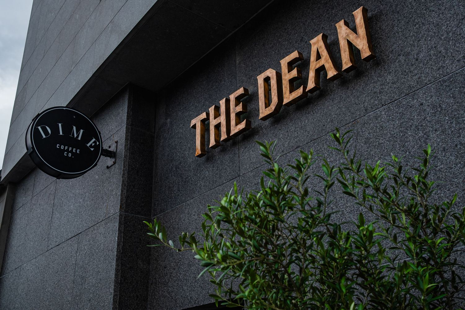 Black and gold restaurant signage mounted on a gray stone wall with green plants underneath.