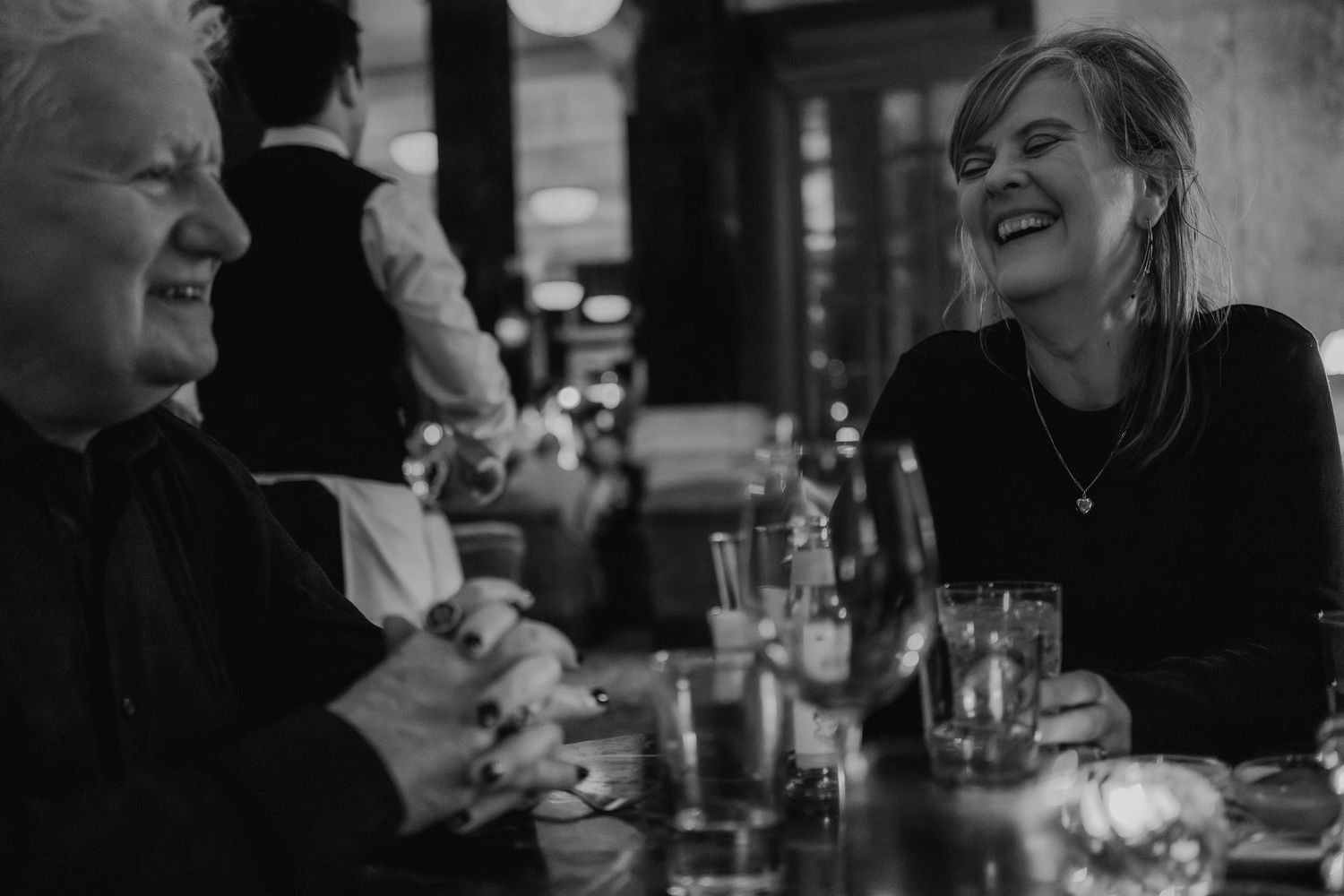 Black and white candid photo of people laughing together at restaurant table.