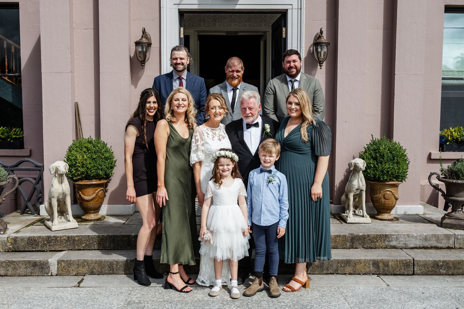 A group photo of wedding guests standing on stone steps with potted plants and statues.