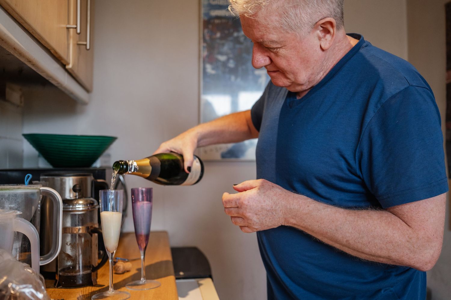 A person in a navy shirt pouring champagne into a glass in a kitchen setting.