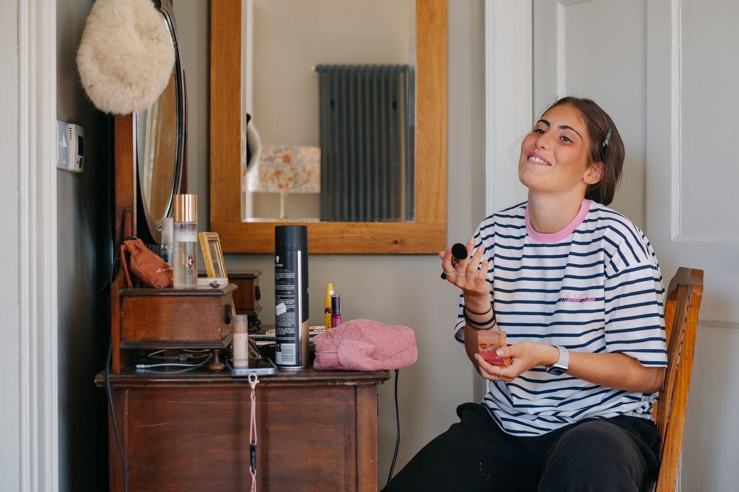 Smiling woman applying makeup with a brush while sitting in a wooden chair near a vintage dressing table.