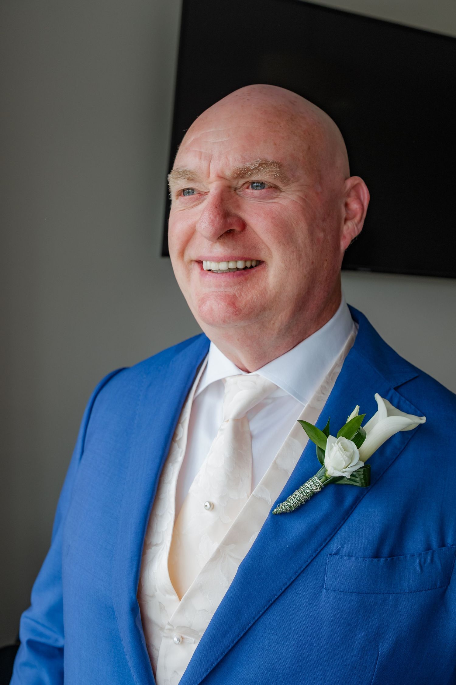 Smiling bald groom in blue suit with white boutonniere and cream waistcoat at wedding.
