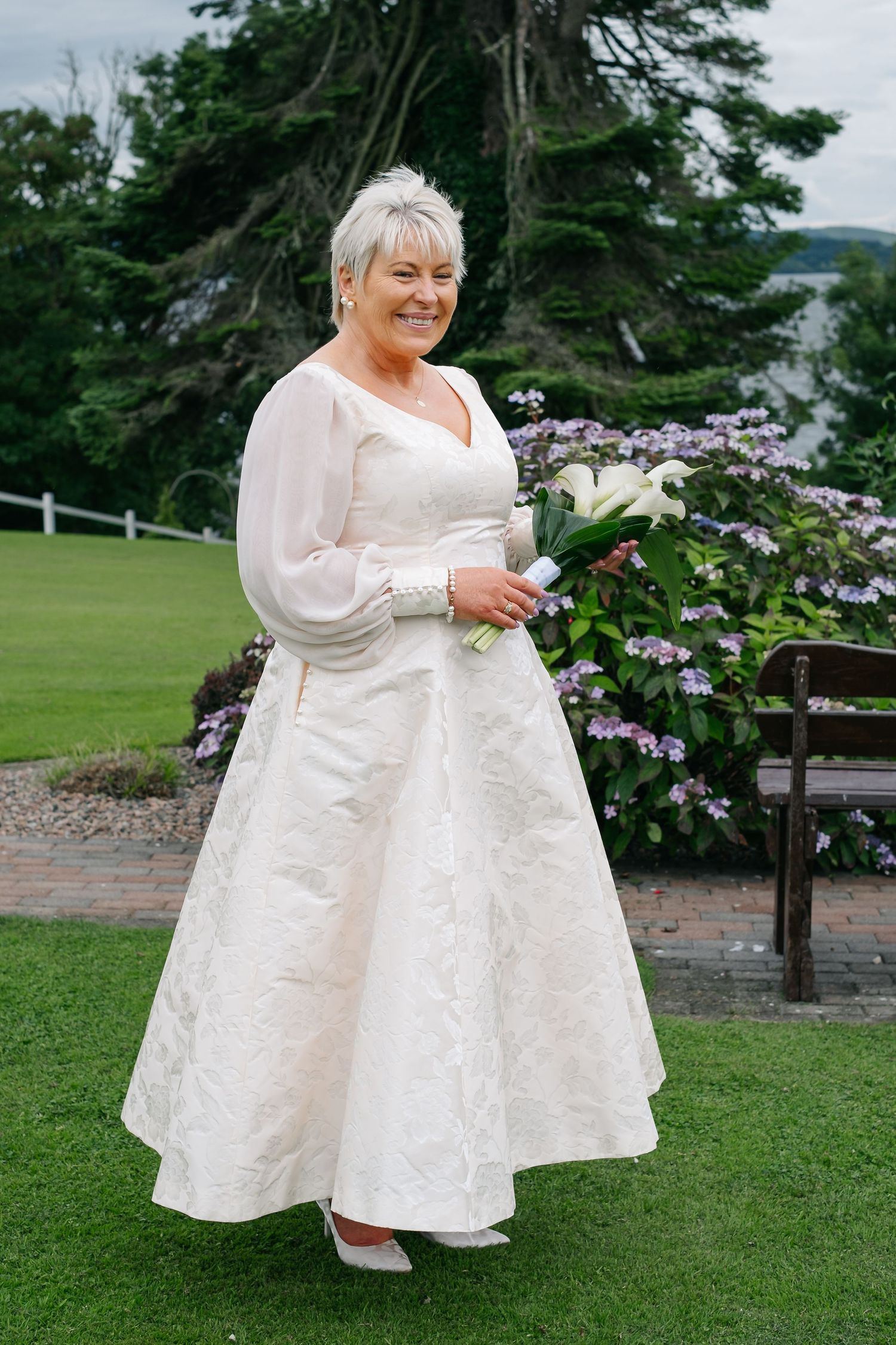 Smiling mature bride in white lace wedding gown holding calla lily bouquet in garden.