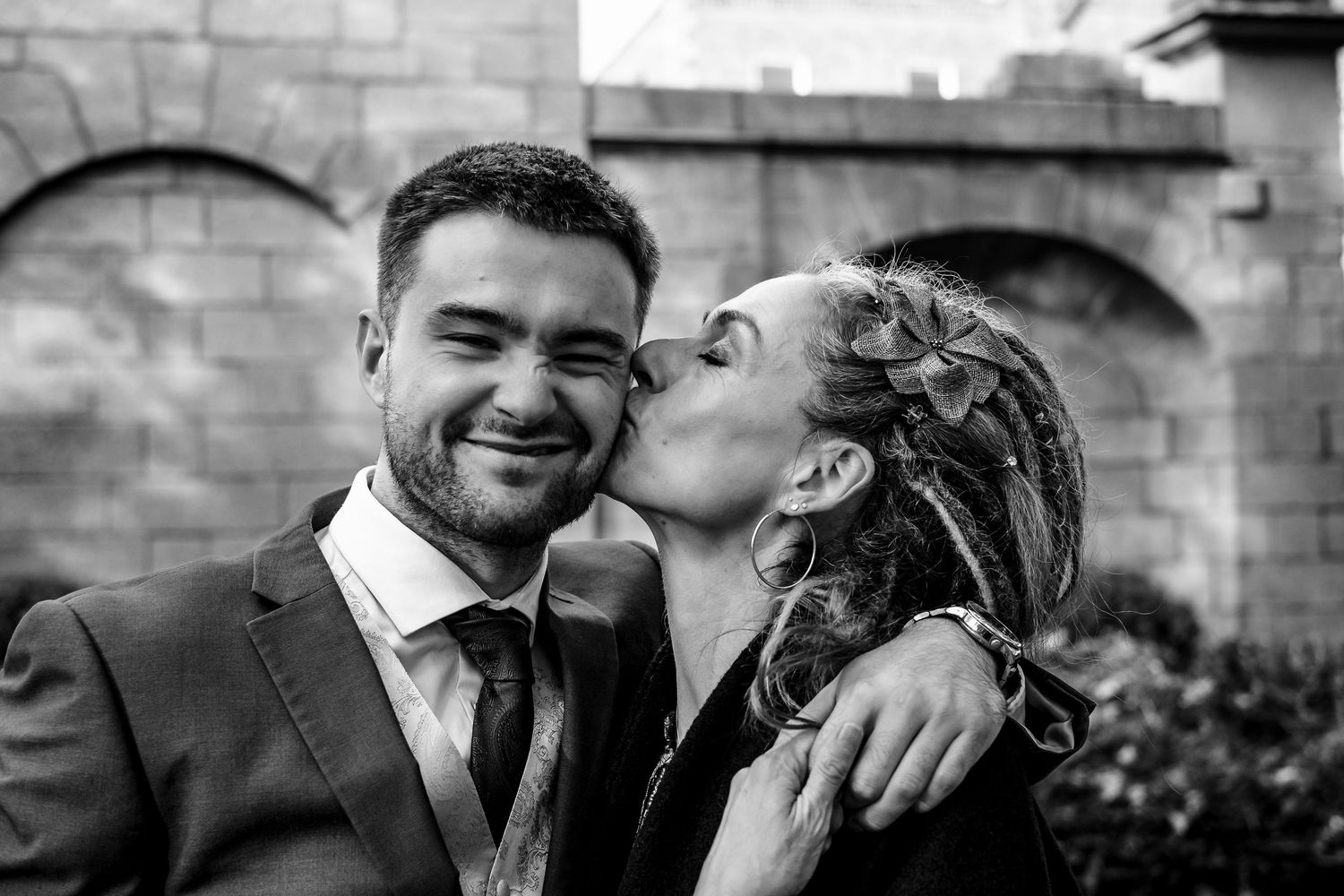Black and white photo of a smiling groom being kissed on the cheek by a woman with dreads at a wedding.