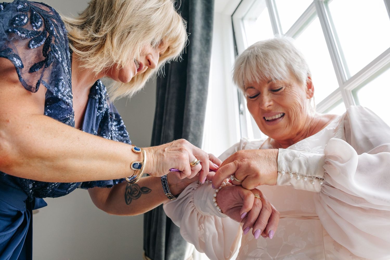 A tattooed woman helps an older bride fasten her white wedding dress sleeve buttons before the ceremony.