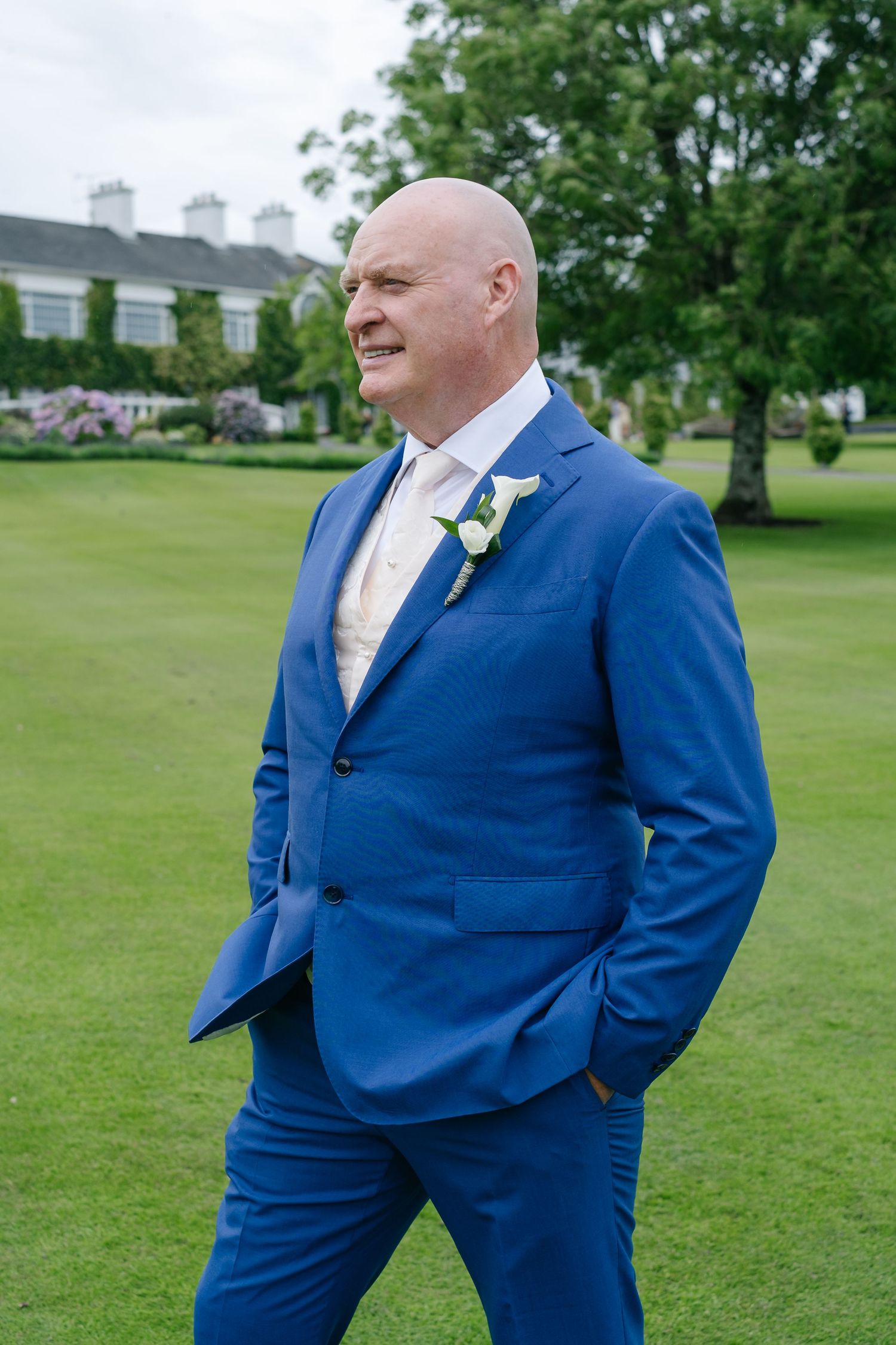Confident bald groom in royal blue suit with white boutonniere walking on green lawn.