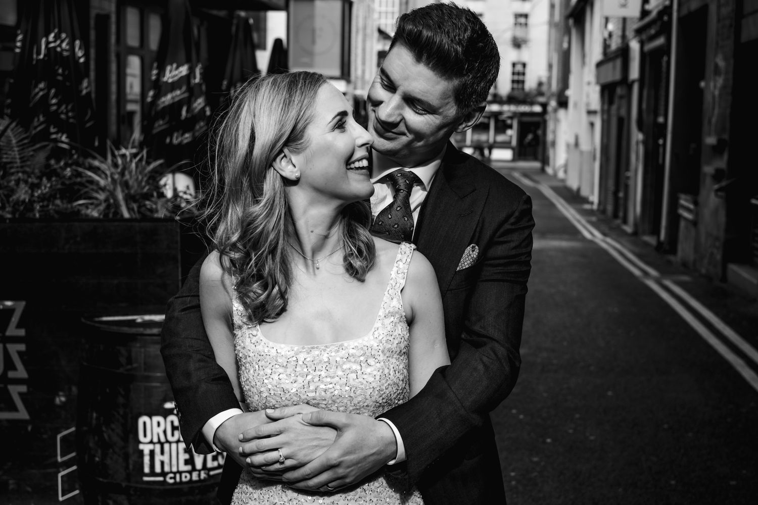 Black and white photo of a smiling couple in wedding attire embracing on a cobblestone street.