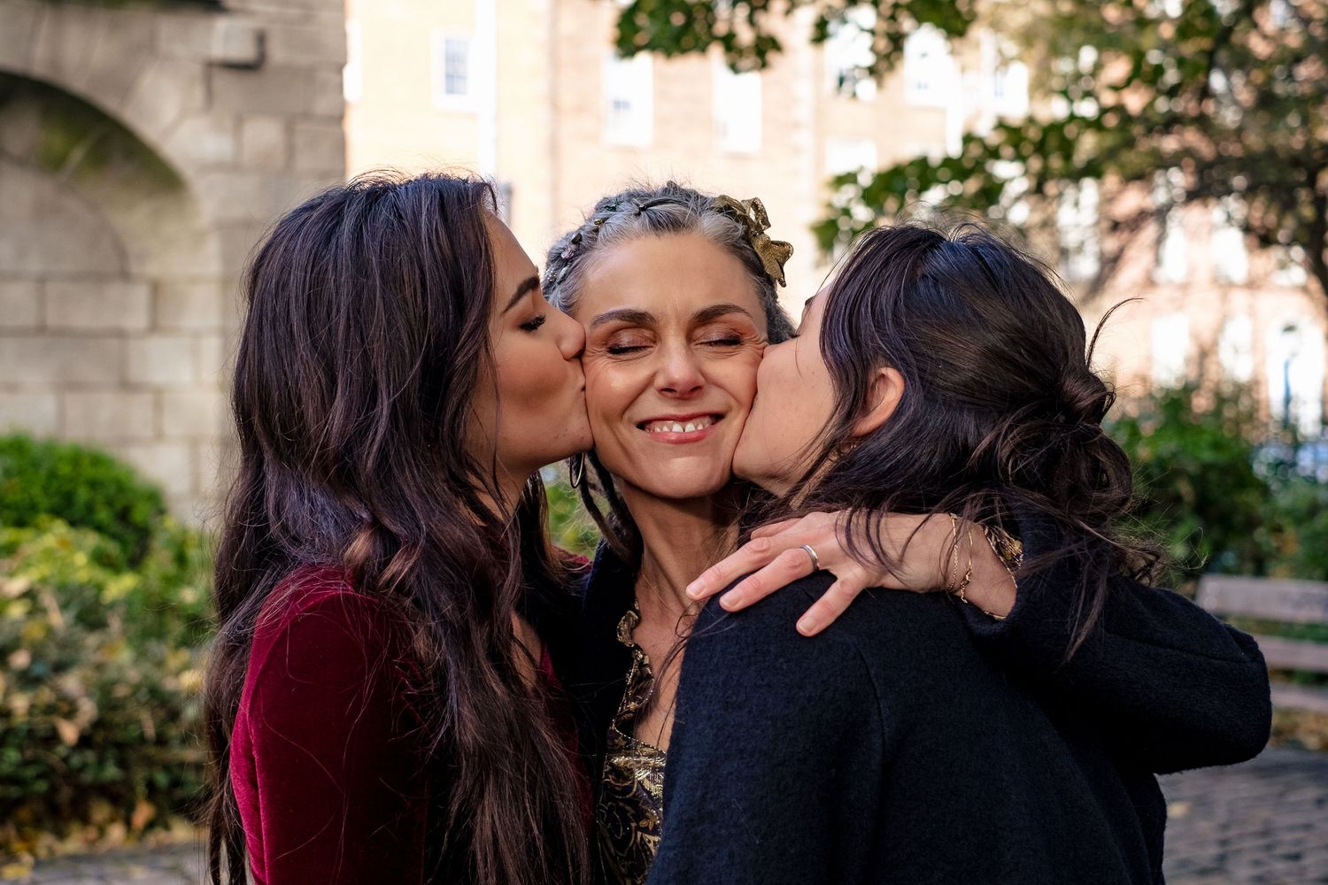 Two women kissing the cheeks of a smiling woman outdoors near a stone building.