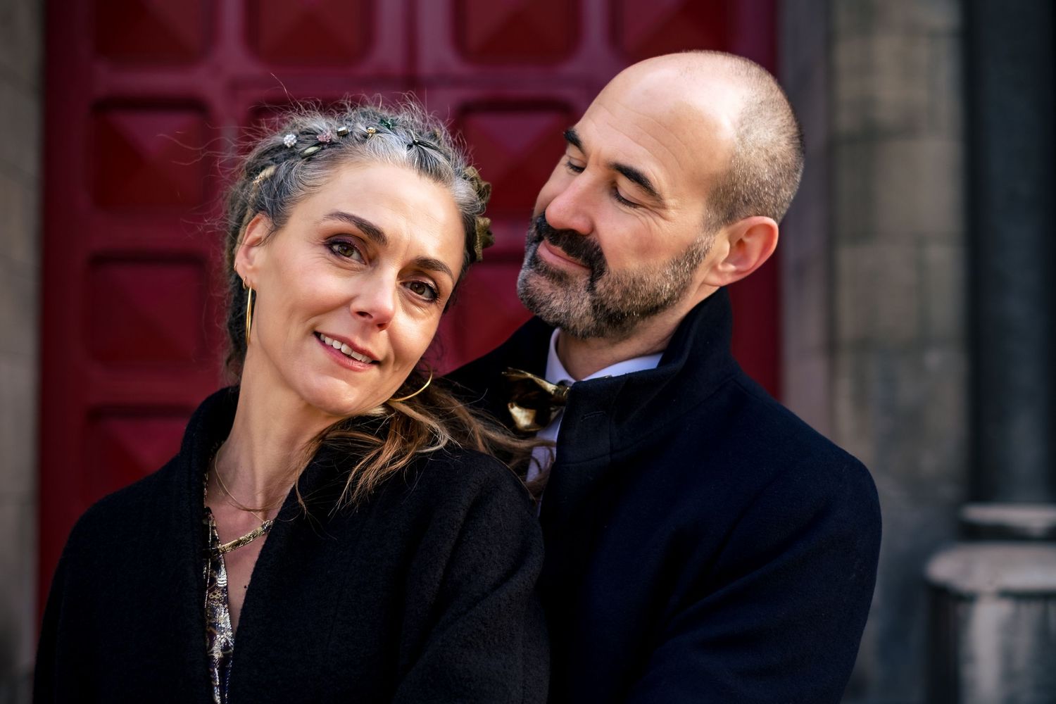 A smiling couple in dark coats poses warmly together in front of a red door.