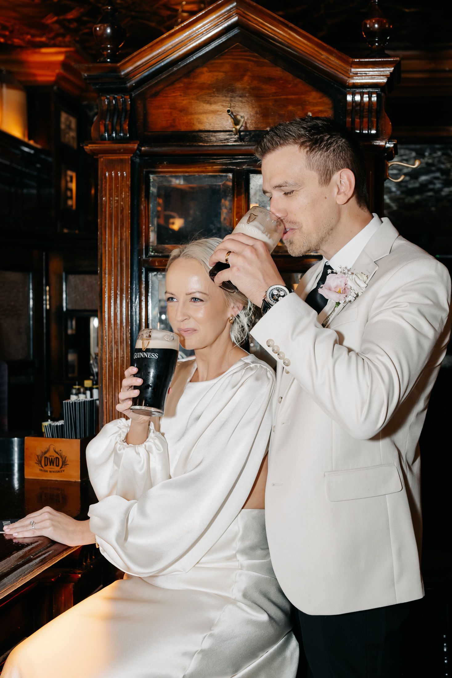 Bride and groom in white attire drinking Guinness pints together at a dark wood bar on their wedding day.
