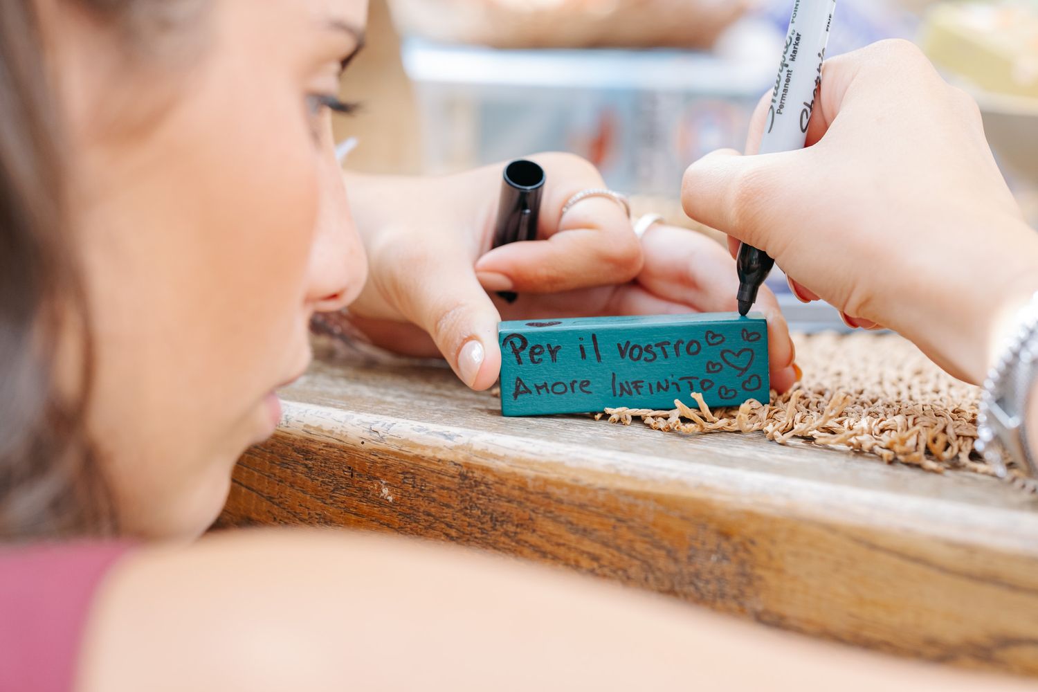 Woman writing Italian text on a teal label with a black marker at a wooden craft table.