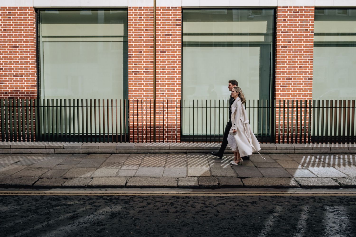 Person in light coat walks past modern storefront with iron railings in urban setting.