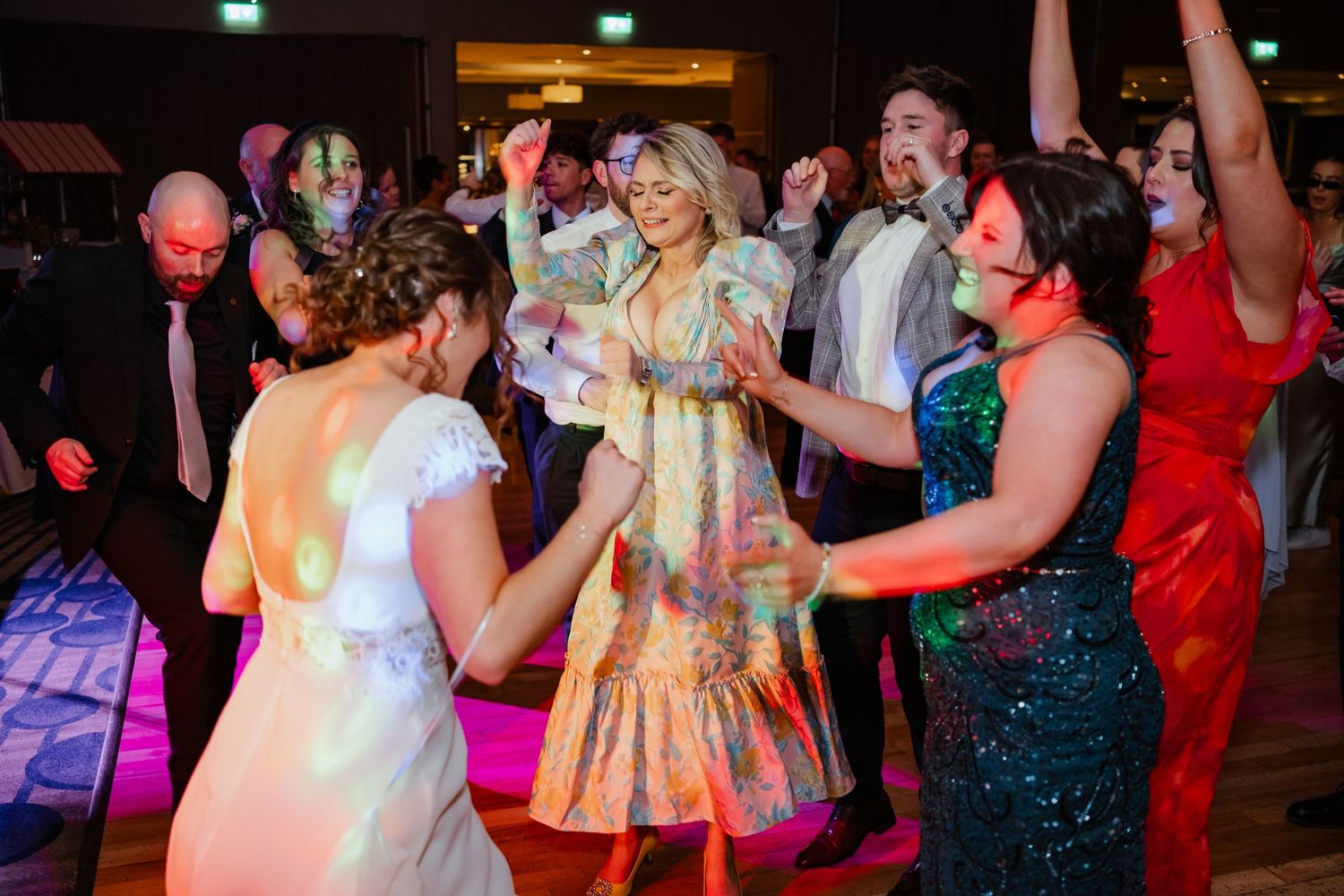 Wedding guests dance together on a crowded dance floor with colorful lighting and festive atmosphere.