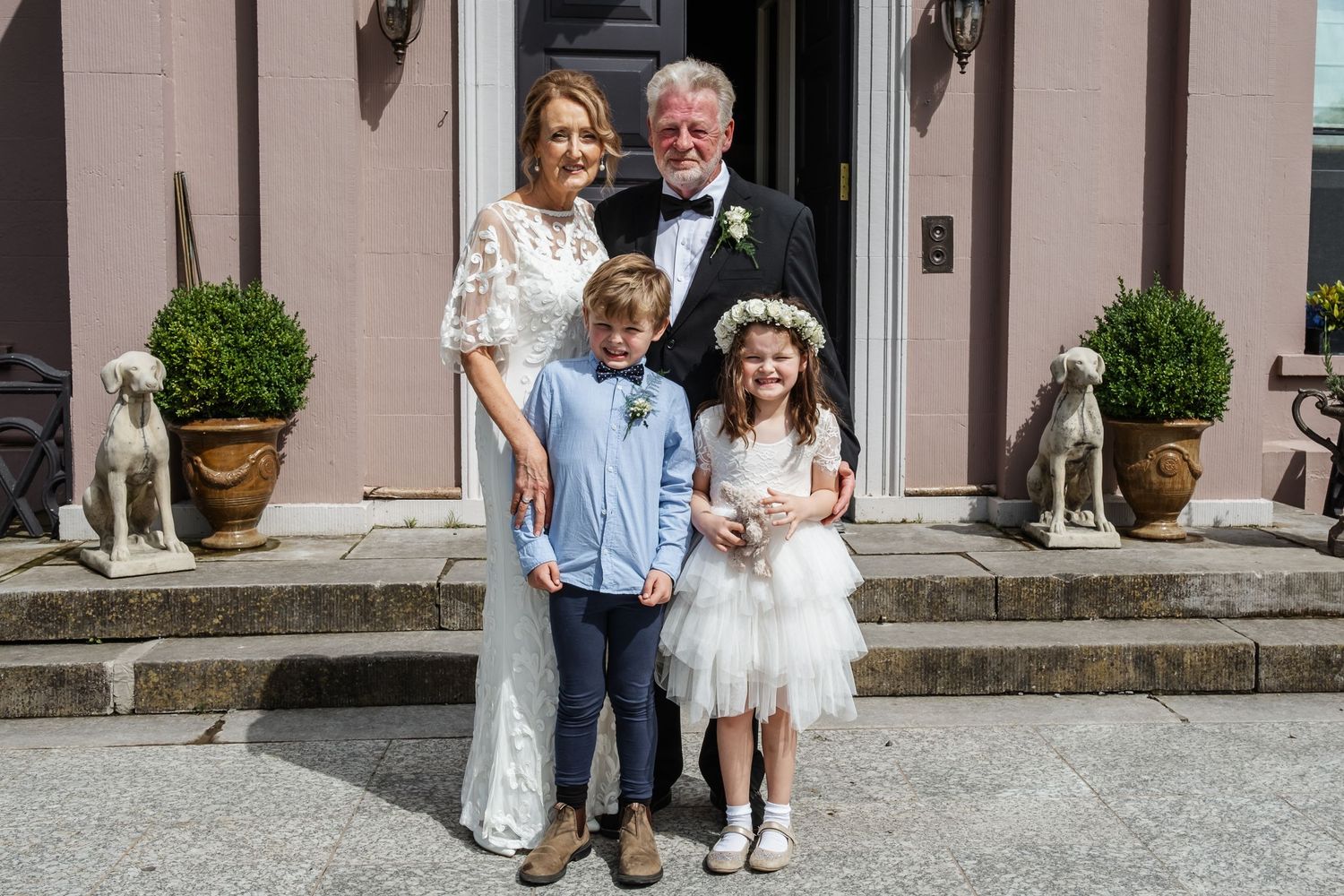 A formal family portrait on stone steps with decorative planters and a white dog.