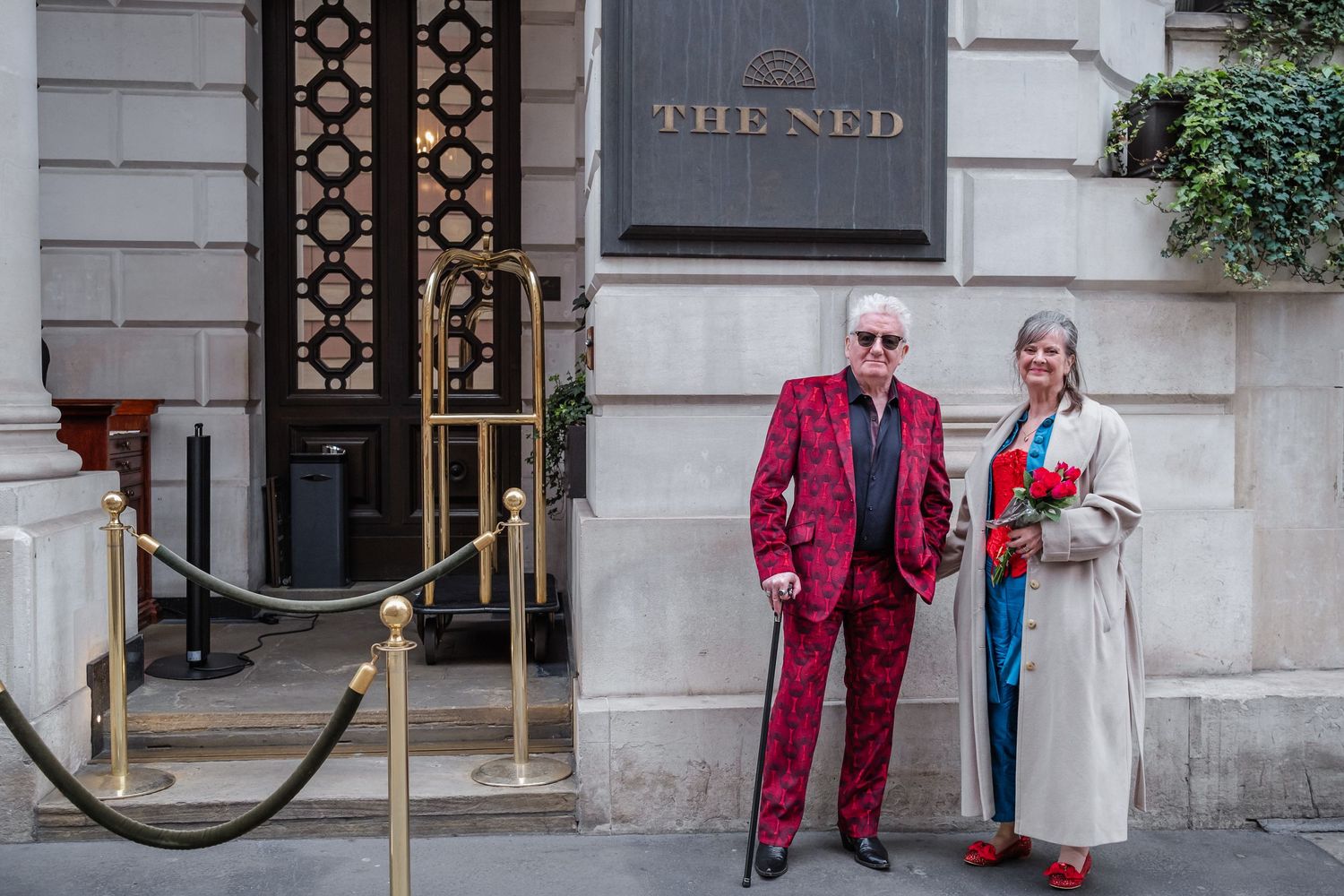 Two people in vibrant outfits pose outside The Ned luxury hotel entrance.