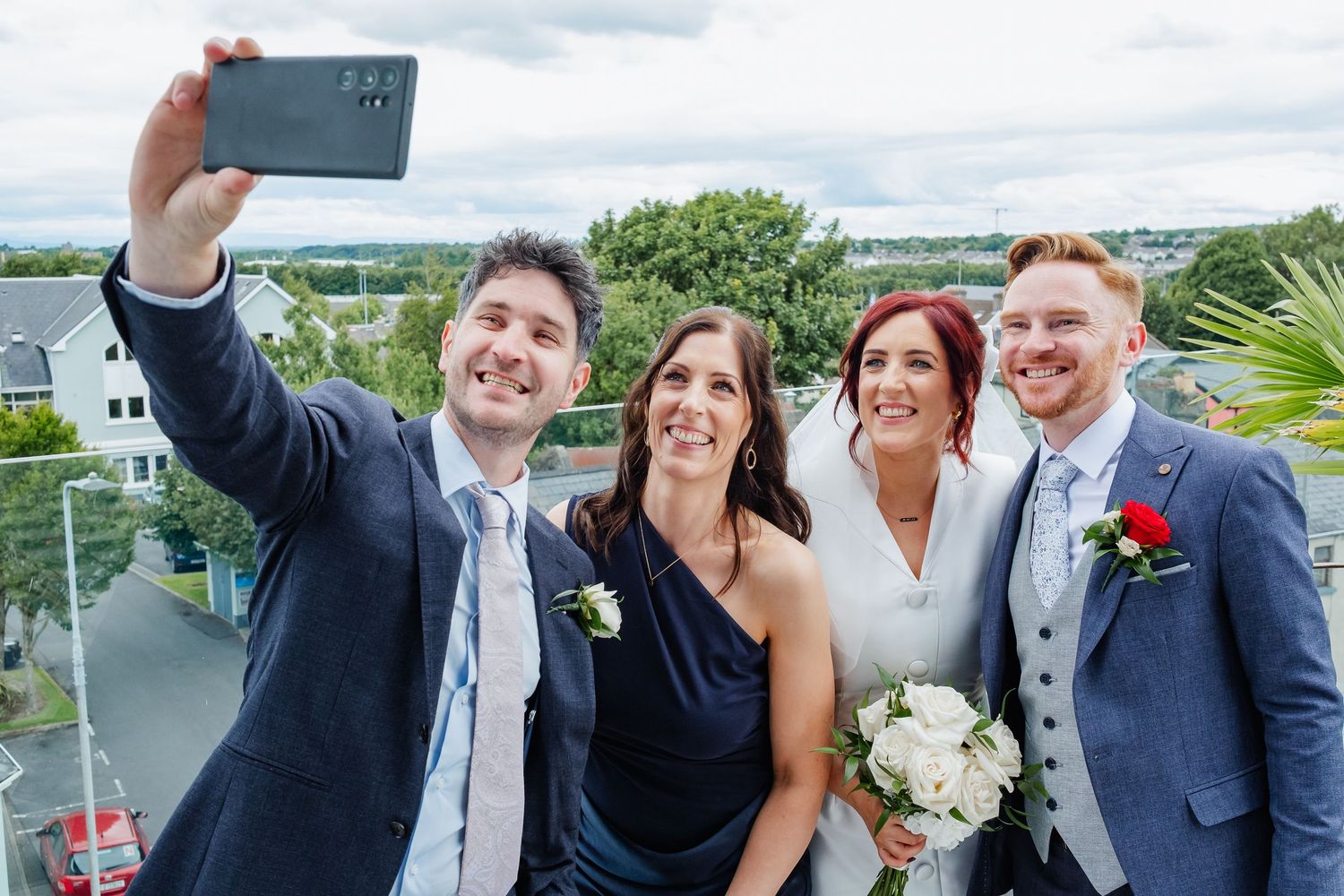 Wedding party taking cheerful selfie on outdoor terrace with cloudy sky backdrop.