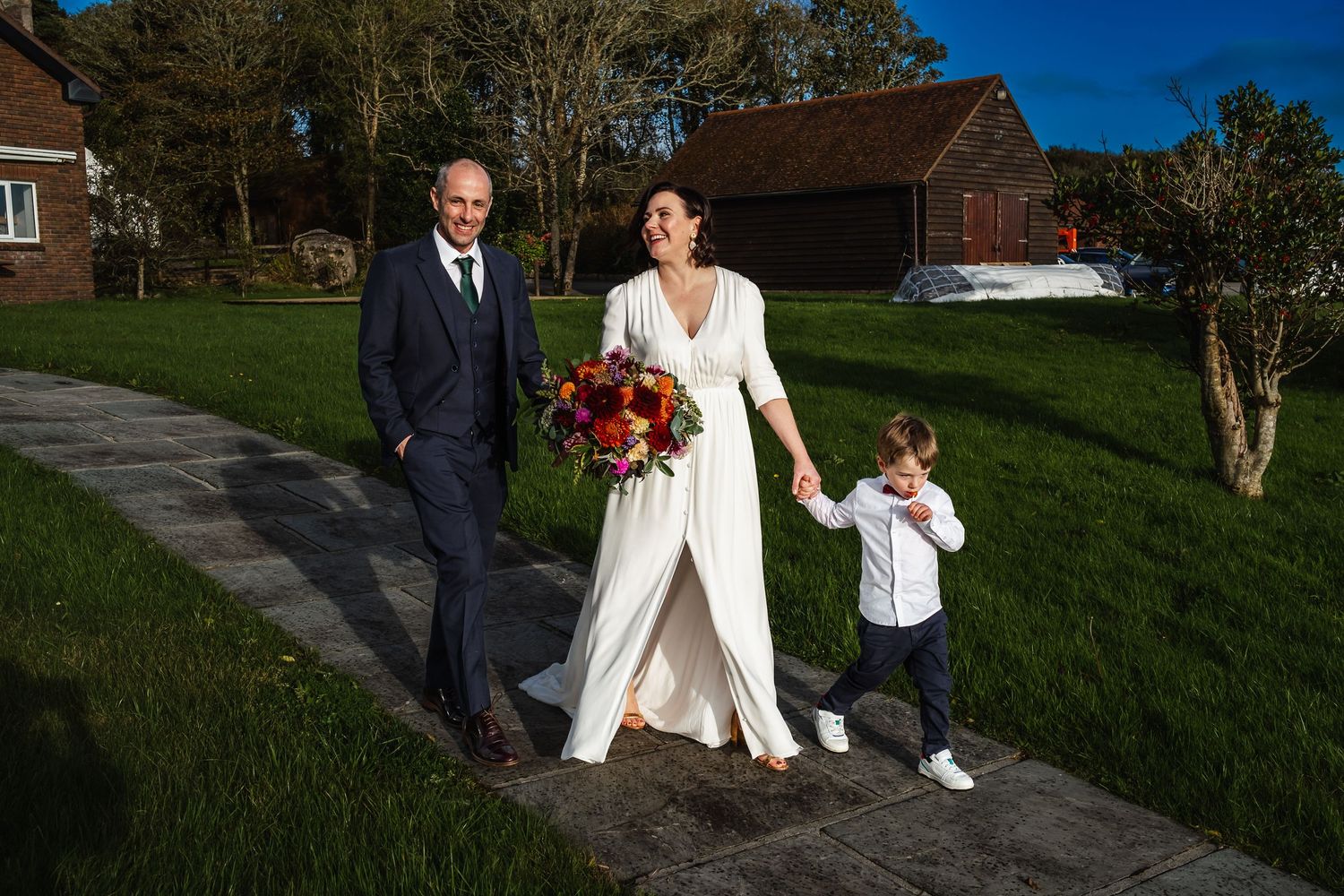 Bride in white gown holds bouquet walking with groom in navy suit and young boy on sunny wedding day.