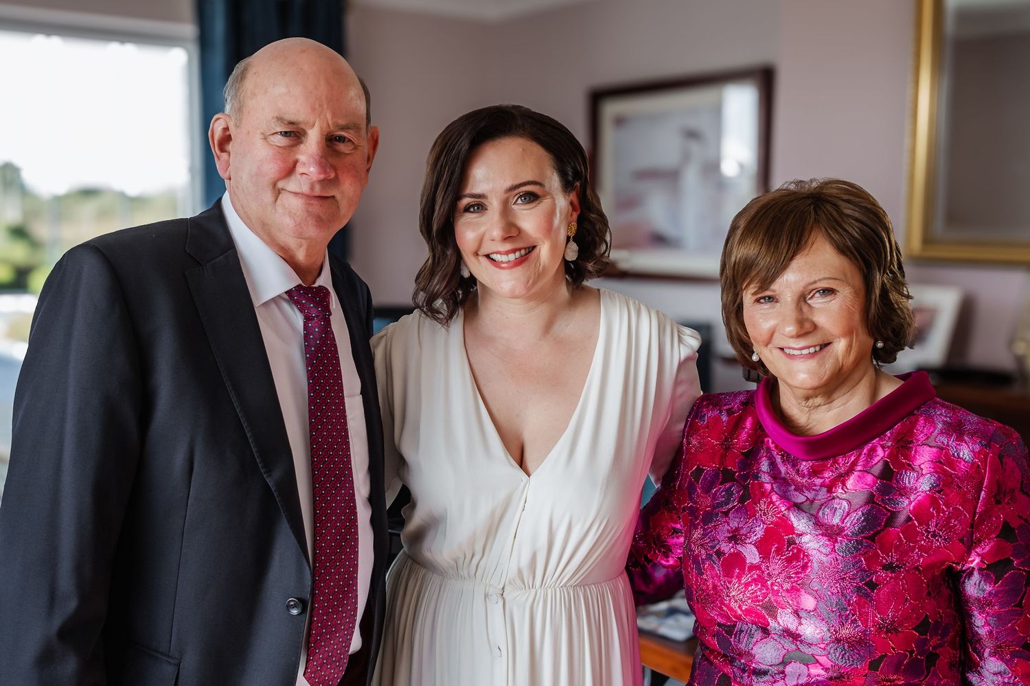 Bride in white dress smiling with two guests at a wedding celebration indoors.