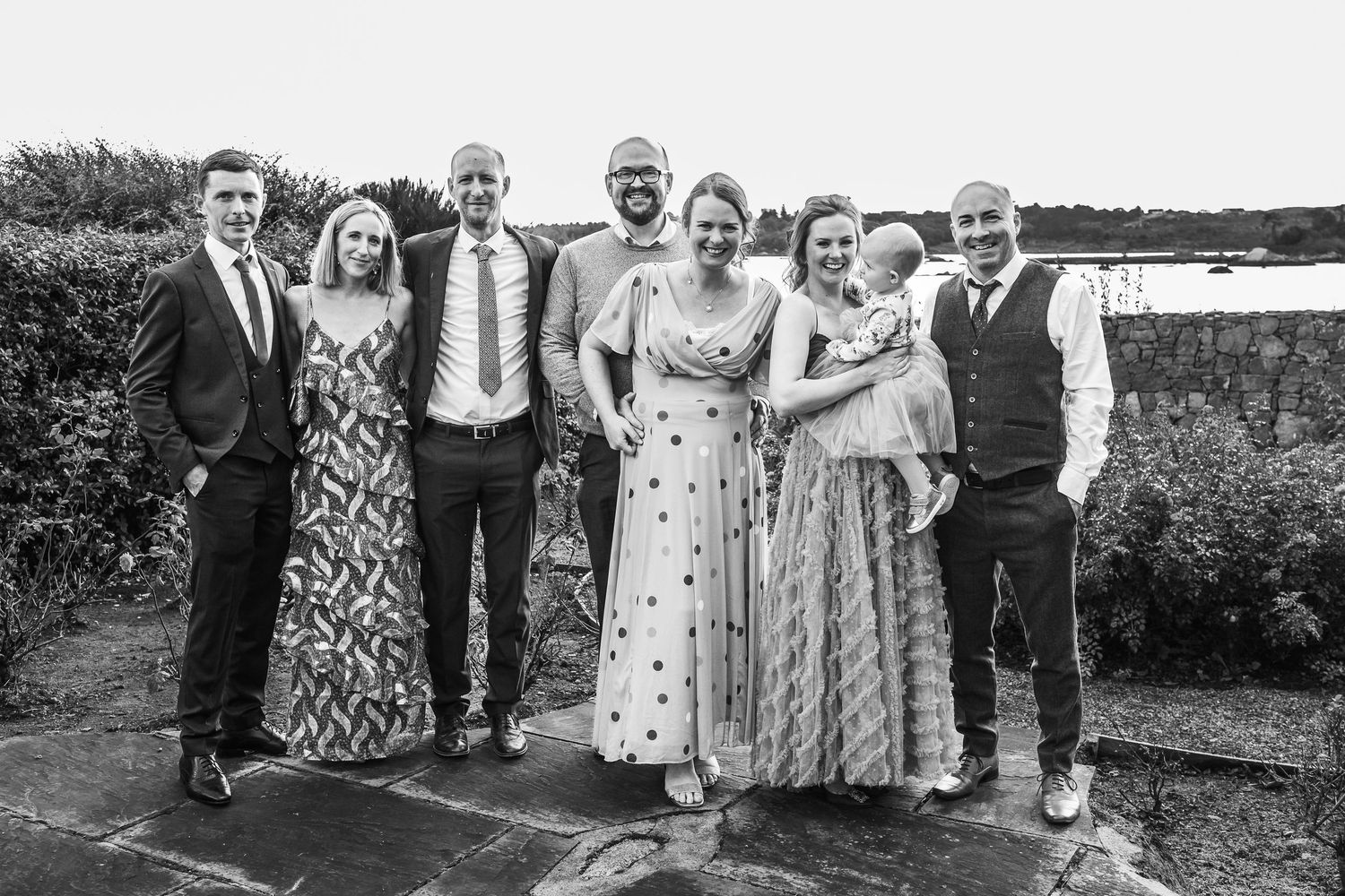 Black and white photo of seven people posing together outdoors at a wedding celebration near a waterfront.