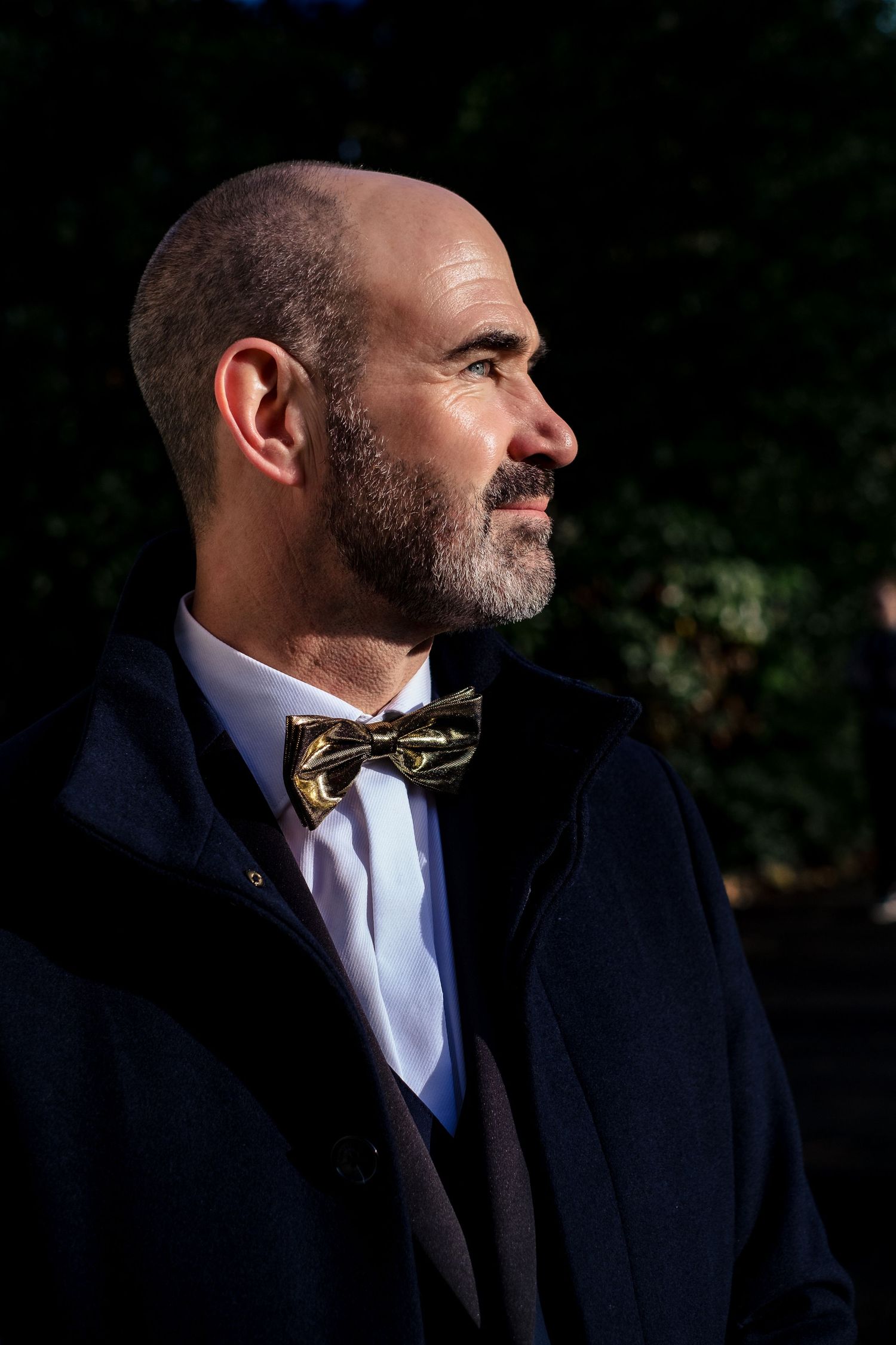 Bald man with beard wearing gold bow tie and navy coat in profile view outdoors.