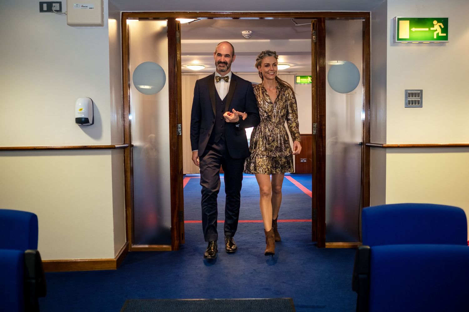 A well-dressed couple walks together through a doorway in a modern corridor with blue chairs.