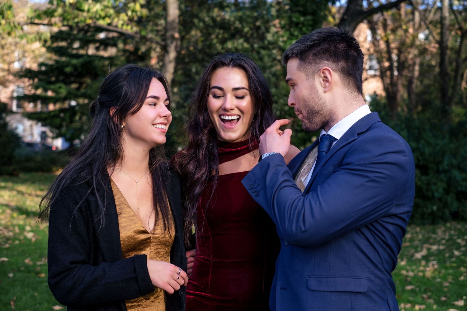 Two women and a man laughing together outdoors in formal attire, posing for a photo in a green park setting.