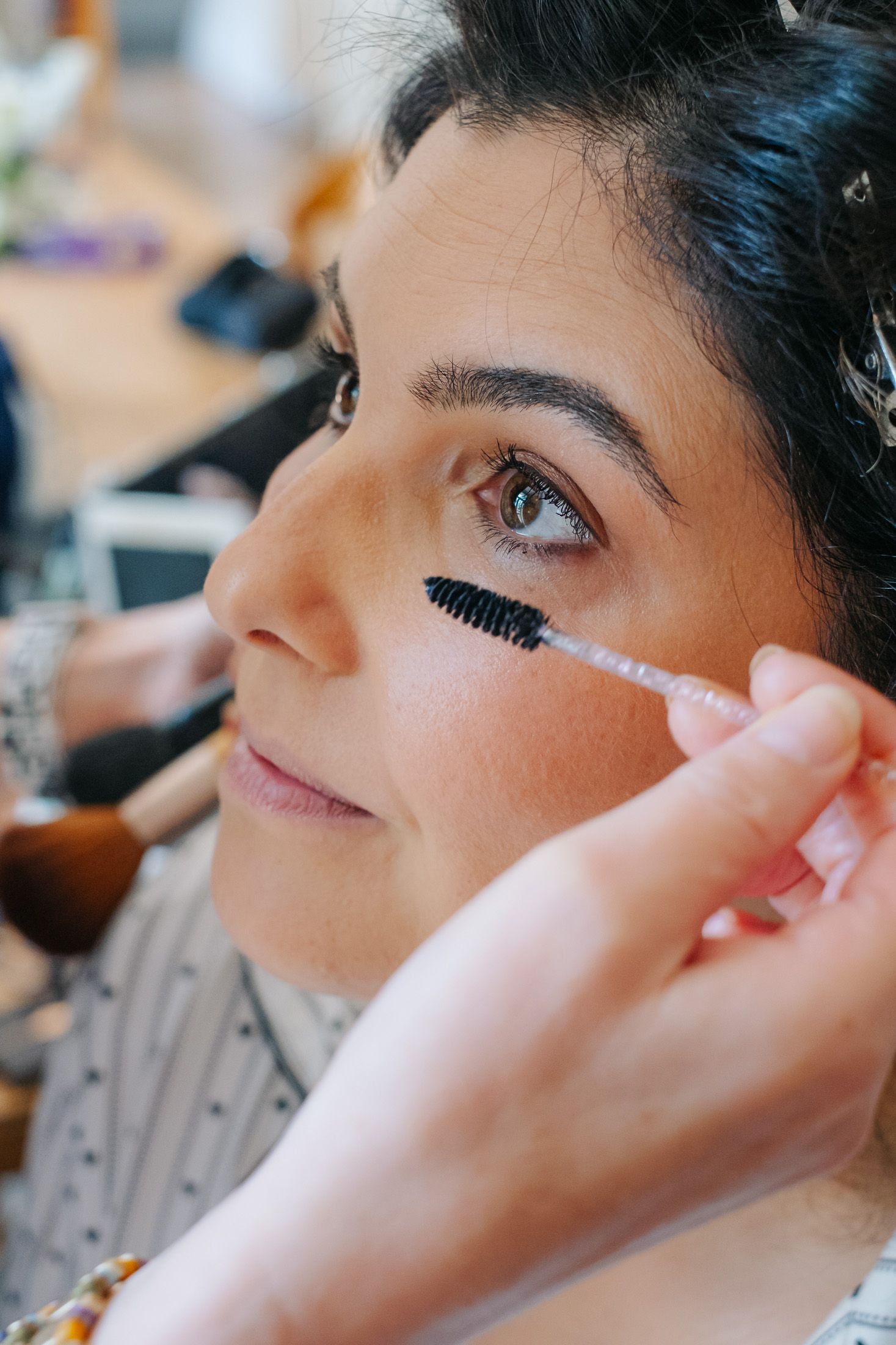 A makeup artist applying mascara to a young brunette woman's lashes in a professional beauty studio.