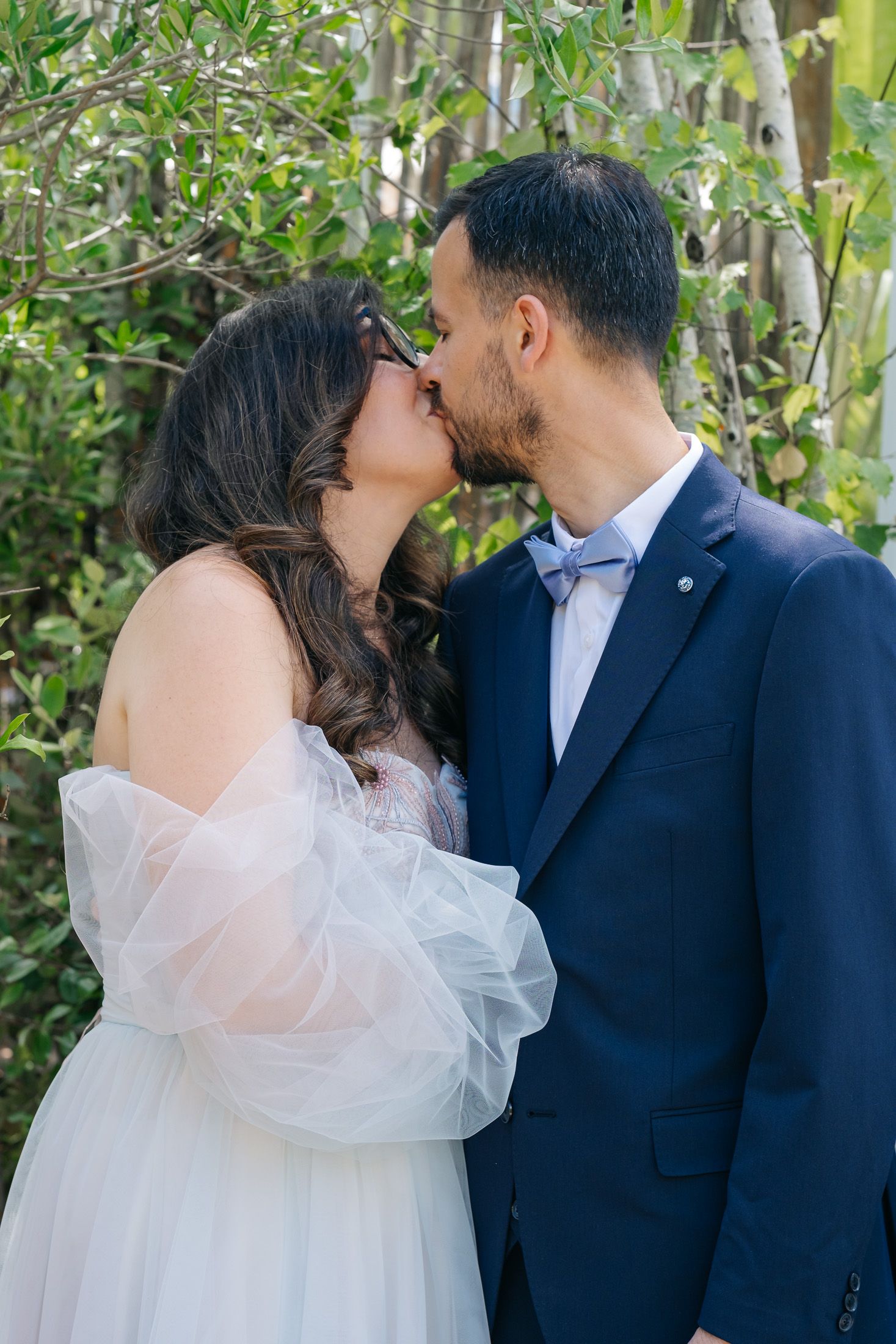 A bride and groom share a romantic kiss outdoors, dressed in a white tulle gown and navy suit with bow tie.