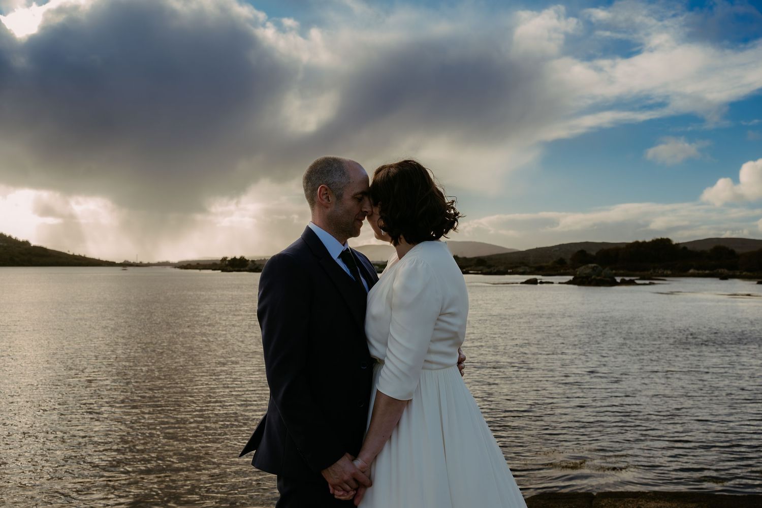 Couple sharing a romantic moment by a lakeside at sunset on their wedding day, with dramatic clouds in the background.