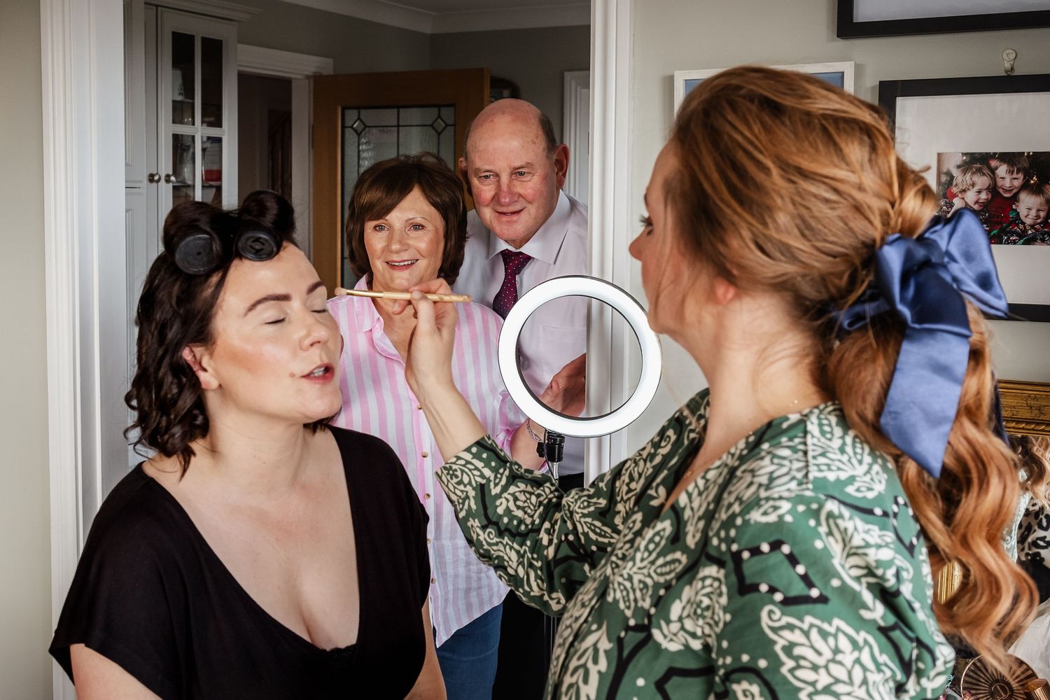 Makeup artist applying makeup to bride while parents watch during wedding morning preparations.
